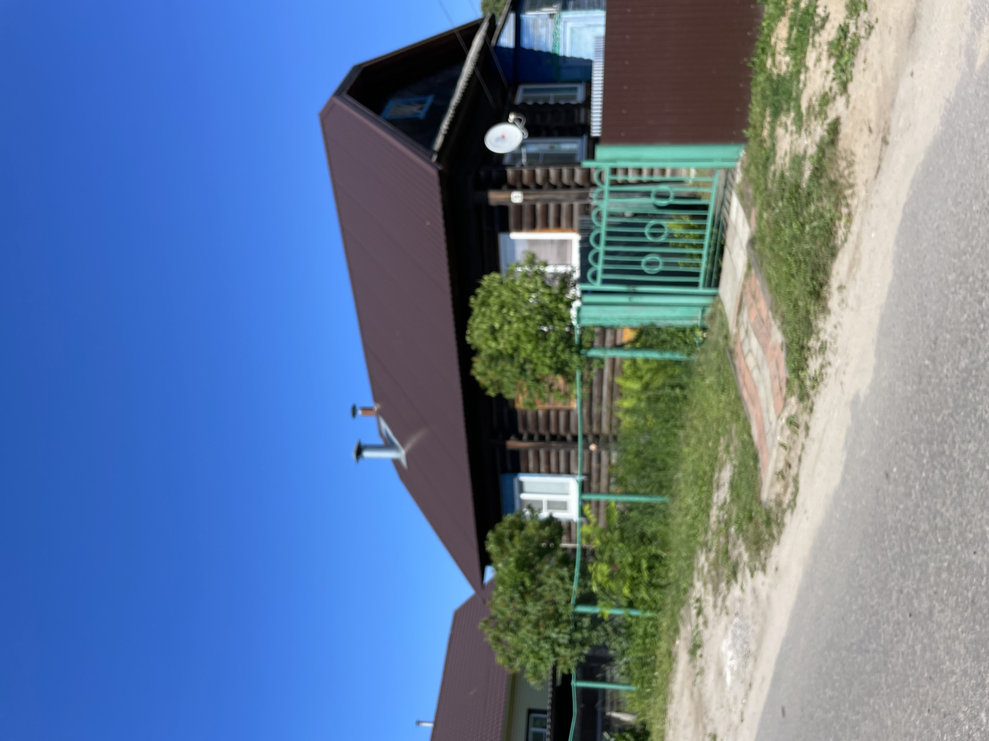 The image captures a residential scene on a sunny day in the village of Krasnye Baki, Russia. In the foreground, a paved road runs horizontally across the bottom of the frame. Beyond the road, a dirt and grass area borders a green metal fence. This fence encloses a property with a prominent dark wooden log house, characterized by a dark brown metal roof and two chimneys. White-framed windows are visible on the house, and a satellite dish is mounted on the exterior wall. To the left of the main house, a second, lighter-colored house with a similar dark brown metal roof is partially visible. Lush green trees and vegetation are present within the fenced area. A green metal gate with circular decorative elements provides access to the property. A brown fence runs along the right side of the property. The sky is a clear, bright blue.