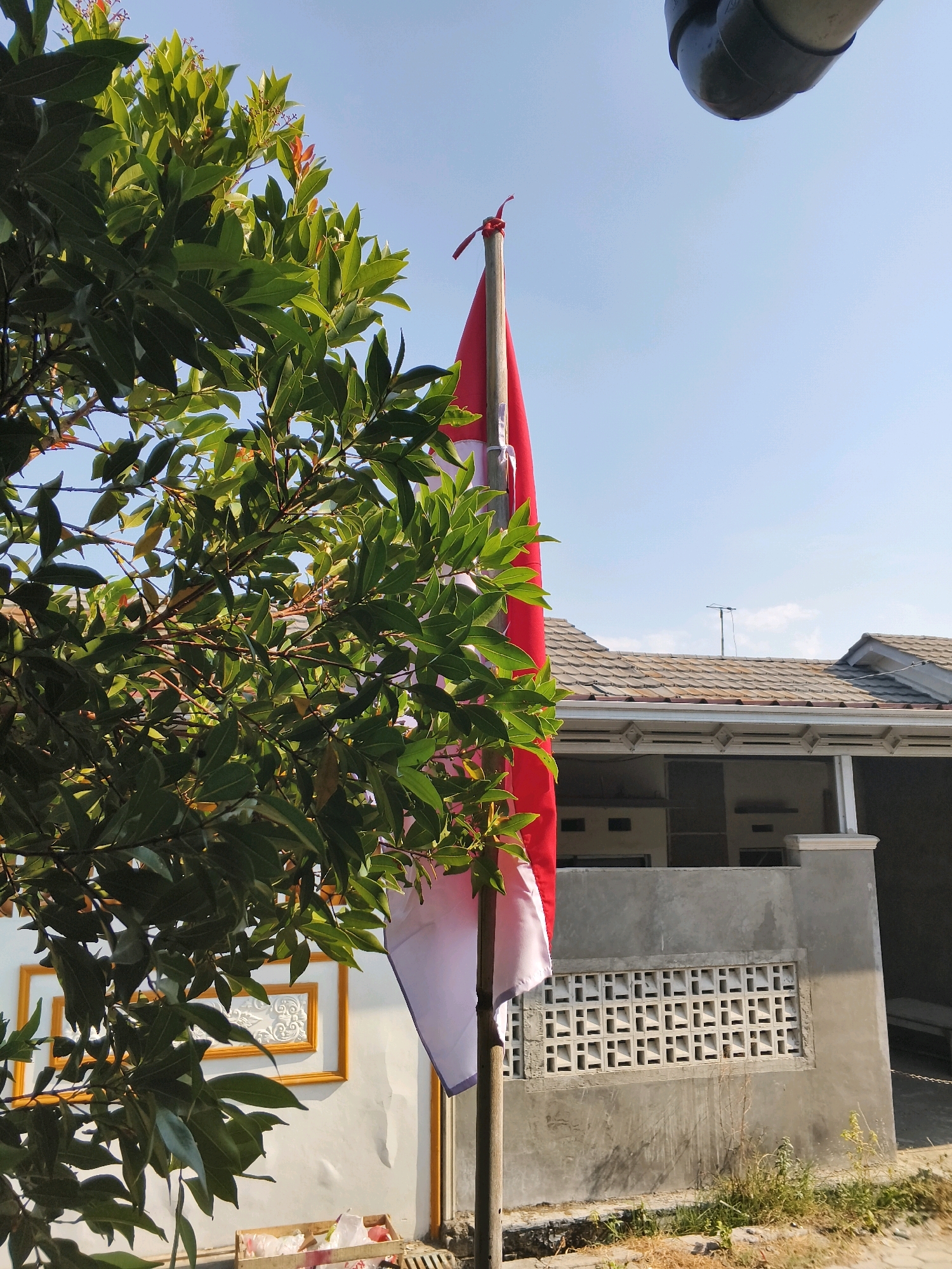 The image shows a red and white flag, likely the Indonesian flag, planted in the ground. A large, leafy green tree is on the left side of the frame, partially obscuring the flag. In the background, there is a building with a tiled roof and a grey concrete wall with a decorative vent. A drainage pipe is visible at the top right of the image. The scene appears to be outdoors, under a clear blue sky, suggesting it is daytime in Bulakelor, Indonesia. There are no people visible in the image.