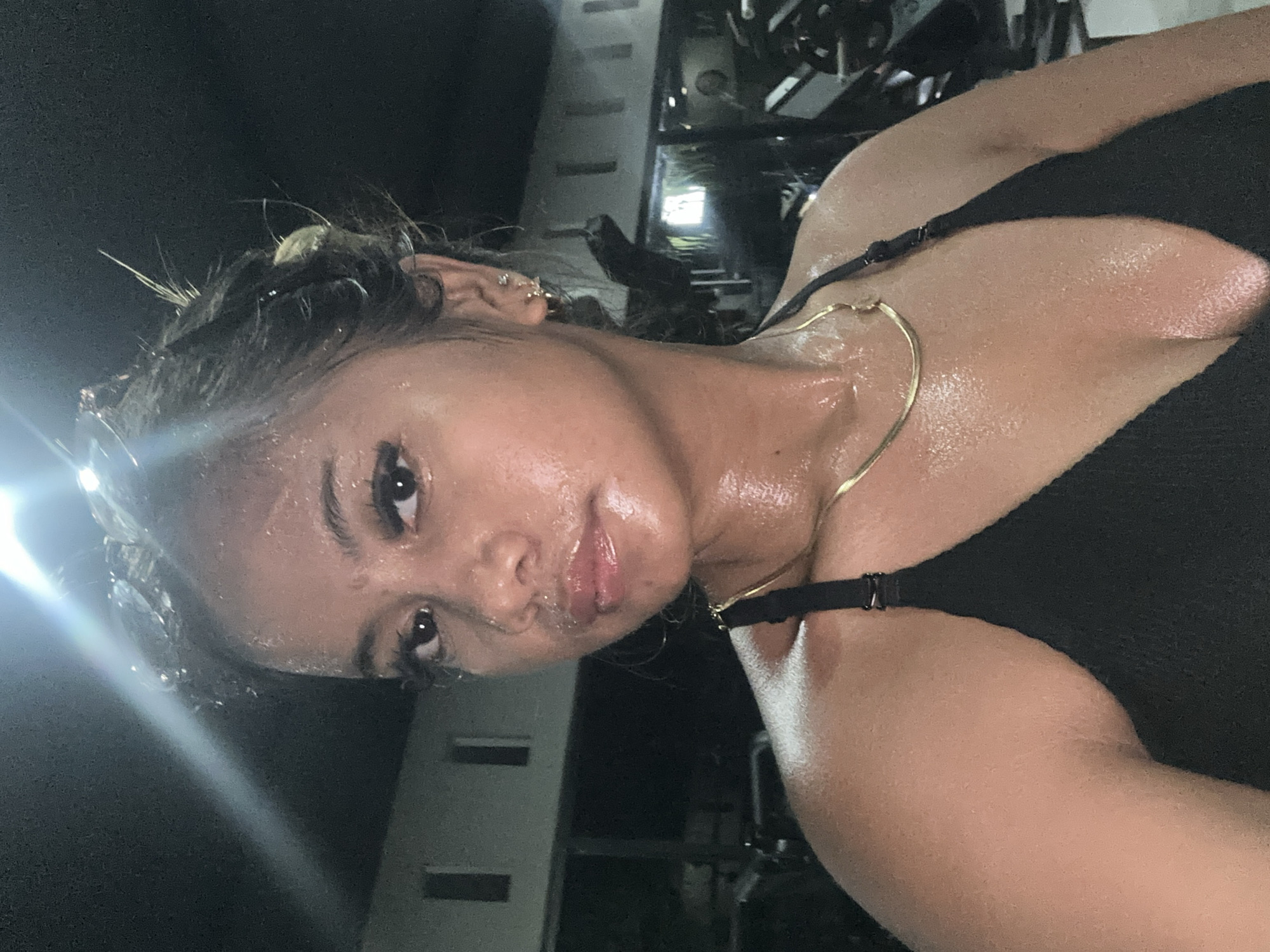 A sweaty woman in a black top with a gold necklace is in the city of Batubulan, Indonesia. The woman is likely at a gym, given the presence of workout equipment in the background. She has dark hair, with some spikes sticking out. The scene is dark, but there is some light from an unknown source.