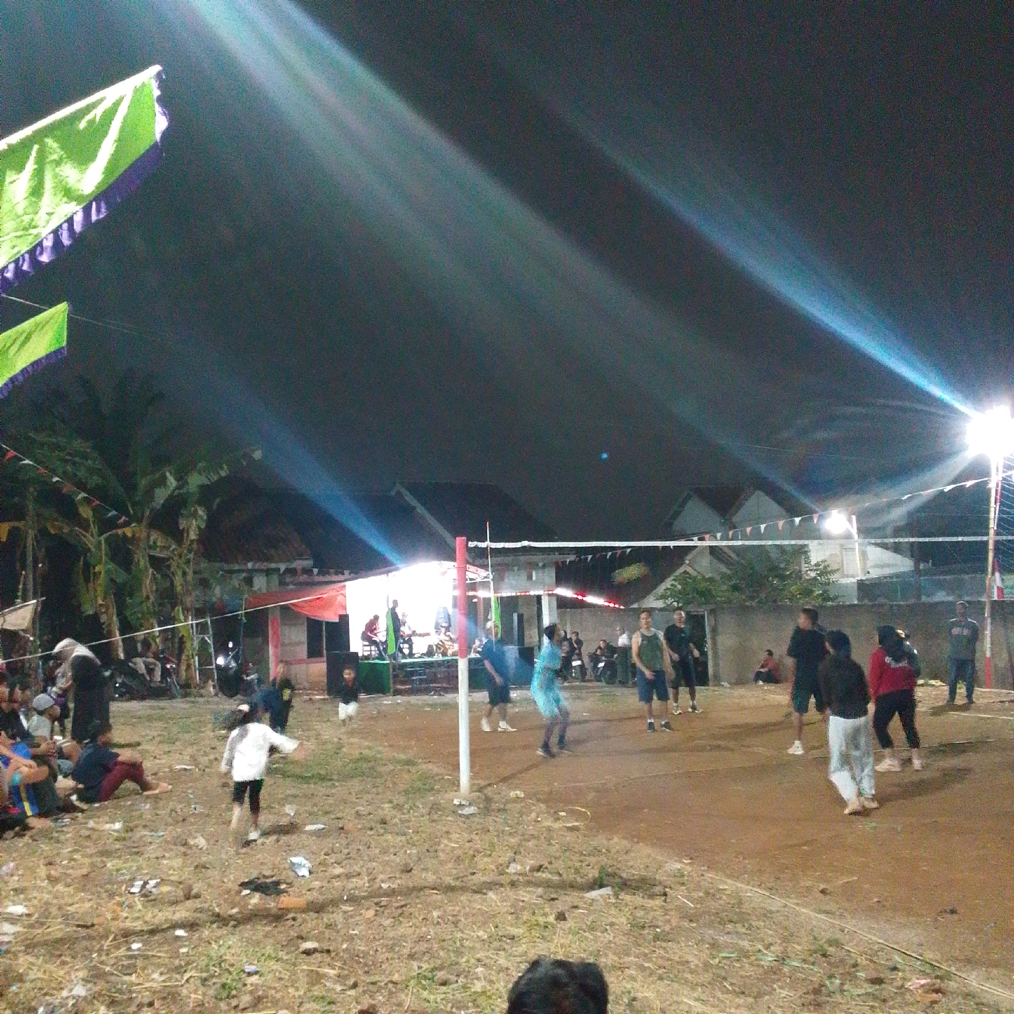 The image depicts an outdoor volleyball game at night, illuminated by artificial lights, in Cilaku, Indonesia. A volleyball net spans the dirt court, with several individuals actively participating in the game. One person in a blue shirt and shorts is in mid-air, seemingly hitting the ball. On the other side of the net, a player in a black shirt and shorts is also positioned to play. Spectators are visible on the periphery of the court, some sitting on the ground and others standing. In the background, there are houses, trees, and tents, suggesting a community event or gathering. Flags and banners are strung across the area, adding to the festive atmosphere. The ground is covered in a mixture of dirt and dry grass, with scattered debris. The lighting creates strong shadows and highlights, casting beams across the scene.