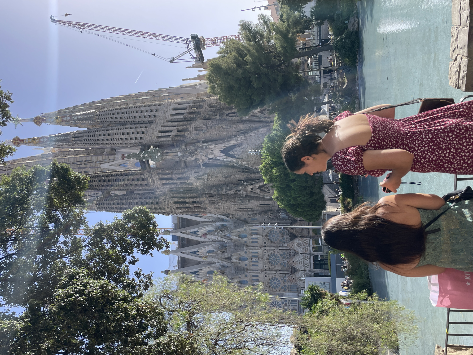 Two young women stand in front of the Sagrada Familia in Barcelona, Spain. The woman on the left has long, dark hair and wears a green strapless dress. She is holding a pink shopping bag. The woman on the right has her hair pulled back in a ponytail and wears a short-sleeved, floral-patterned dress. She is holding a small black camera. The Sagrada Familia, an unfinished Roman Catholic minor basilica designed by Antoni Gaudí, is visible in the background. A construction crane is also present, indicating ongoing work on the building. In the foreground, a body of turquoise water reflects the sky and the surrounding trees. The scene is bathed in sunlight, creating lens flare in the upper right portion of the image.