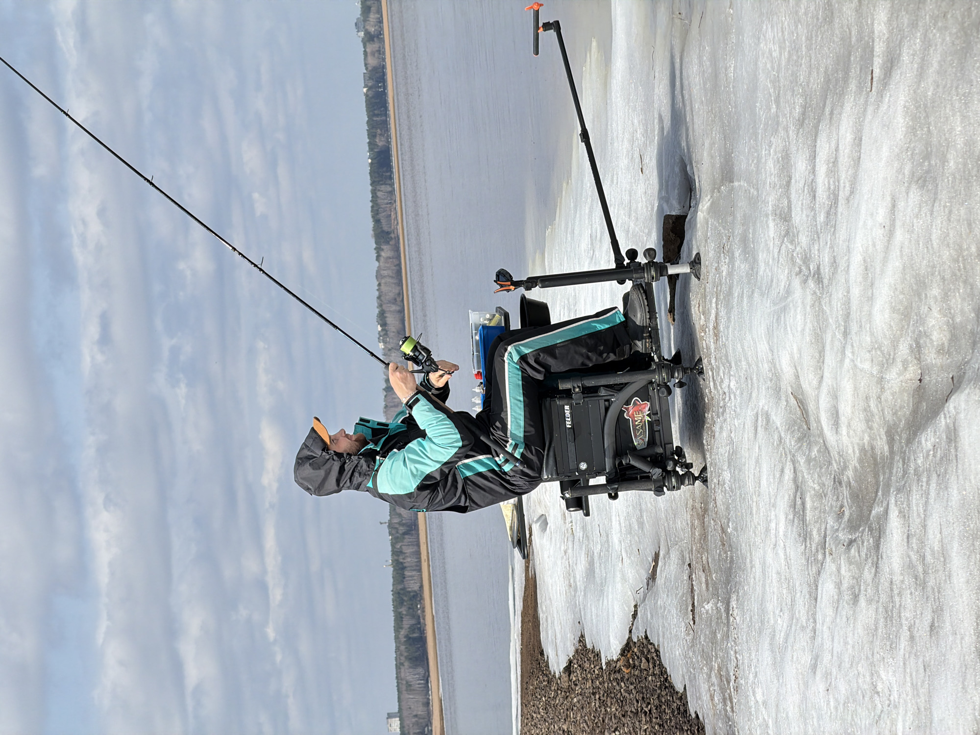 A man is seated on a specialized fishing platform, holding a fishing rod and reel. He is on the ice next to the water. He is wearing a black jacket with mint-colored accents and a hood. The platform has the word "FEEDER" on it, and a logo with a fish and the word "SASAME." The ice is dirty and slushy. In the background, there is a shoreline with trees and buildings. The sky is cloudy. The location is Chaykovsky, Russia.