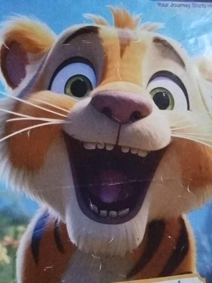 A cartoon tiger with wide green eyes and a joyful, open-mouthed expression dominates the image. It features light orange and cream fur with darker orange markings. A pink nose and a full set of teeth are prominently displayed, suggesting laughter. The tiger's whiskers are thin and white, extending from its cheeks. The background is blurred, giving a hint of greenery and blue, which suggests an outdoor setting.