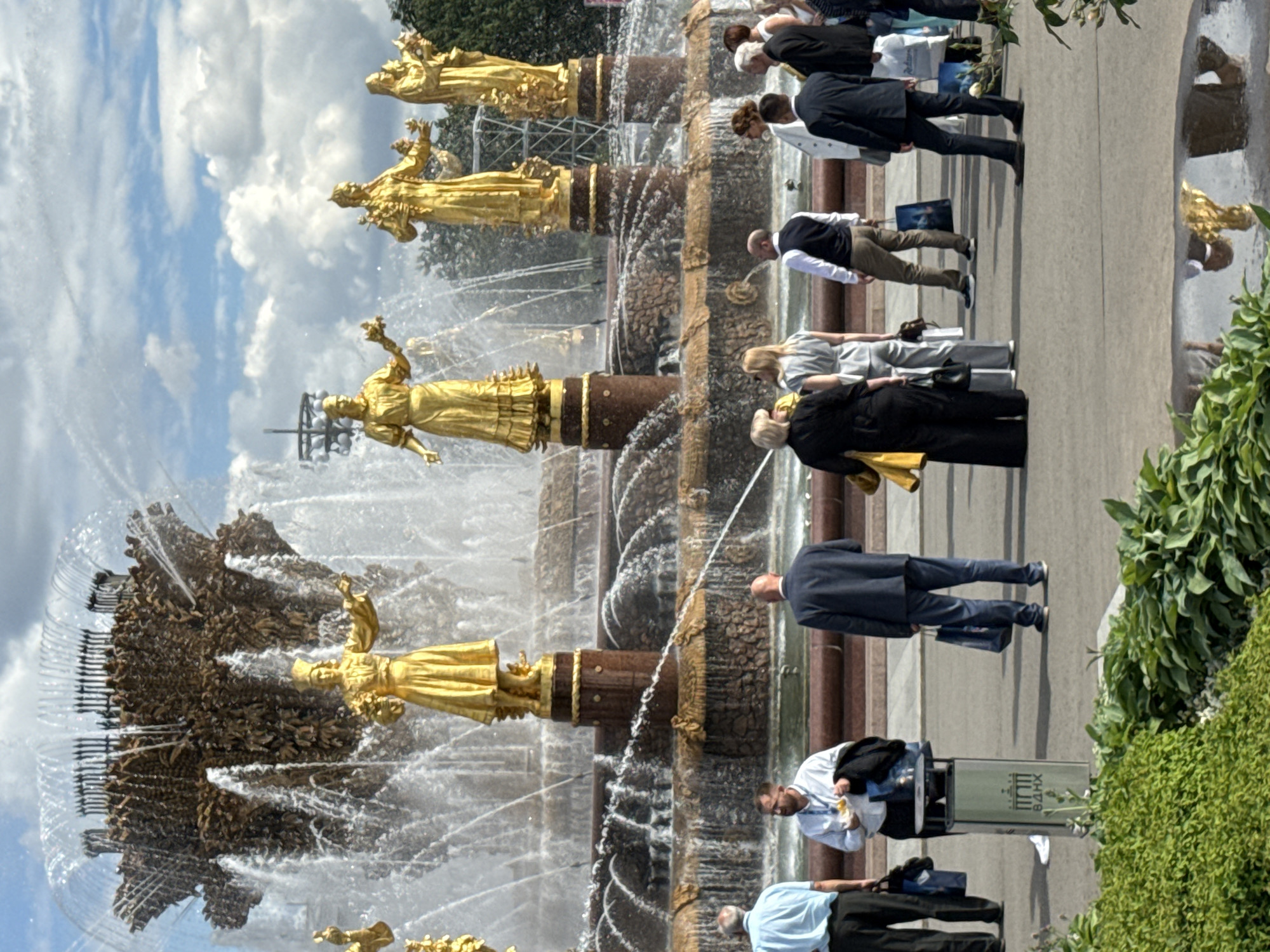 This media file captures a scene at a fountain, likely in Moscow, Russia. The main subjects are several gilded statues of women adorning the tiered fountain, with water spraying from them and around the structure. Surrounding the fountain are numerous people, mostly viewed from behind as they walk or stand, observing the spectacle.
The activity is people enjoying the fountain on what appears to be a bright, sunny day, judging by the shadows and the clear sky with some clouds. The overall mood seems relaxed and leisurely. The statues themselves are impressive, showcasing classical or allegorical figures in dynamic poses. The water creates a sense of movement and freshness.
Notable details include the people's attire, which ranges from casual to more formal wear, suggesting a public space visited by various individuals. Some are dressed in suits, while others are in more casual clothing. There is a trash bin visible with Cyrillic text on it, and the letters "ВДНХ" are clearly discernible. This is a strong indicator that the location is VDNKh (Exhibition of Achievements of National Economy) in Moscow, a well-known park featuring numerous fountains and pavilions. The time of day appears to be daytime, likely late morning or afternoon, given the sunlight.