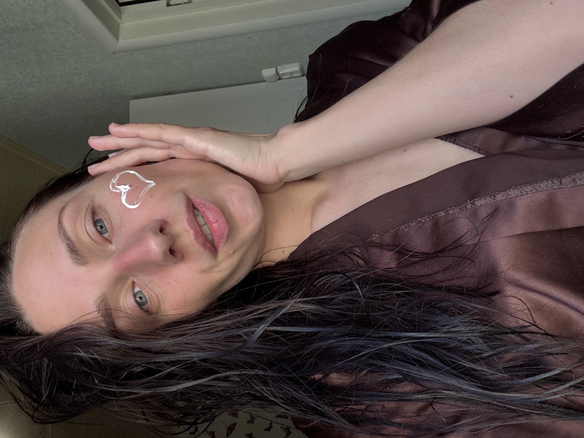 A woman is lying down with her head tilted towards the camera. She has dark, wet hair and is wearing a brown robe. There is a heart-shaped dollop of white cream on her cheek. Her hand is resting on her face. The background is blurry and indistinct, showing a window and part of a wall. There is no indication that this image was taken in Saint Petersburg, Russia.
