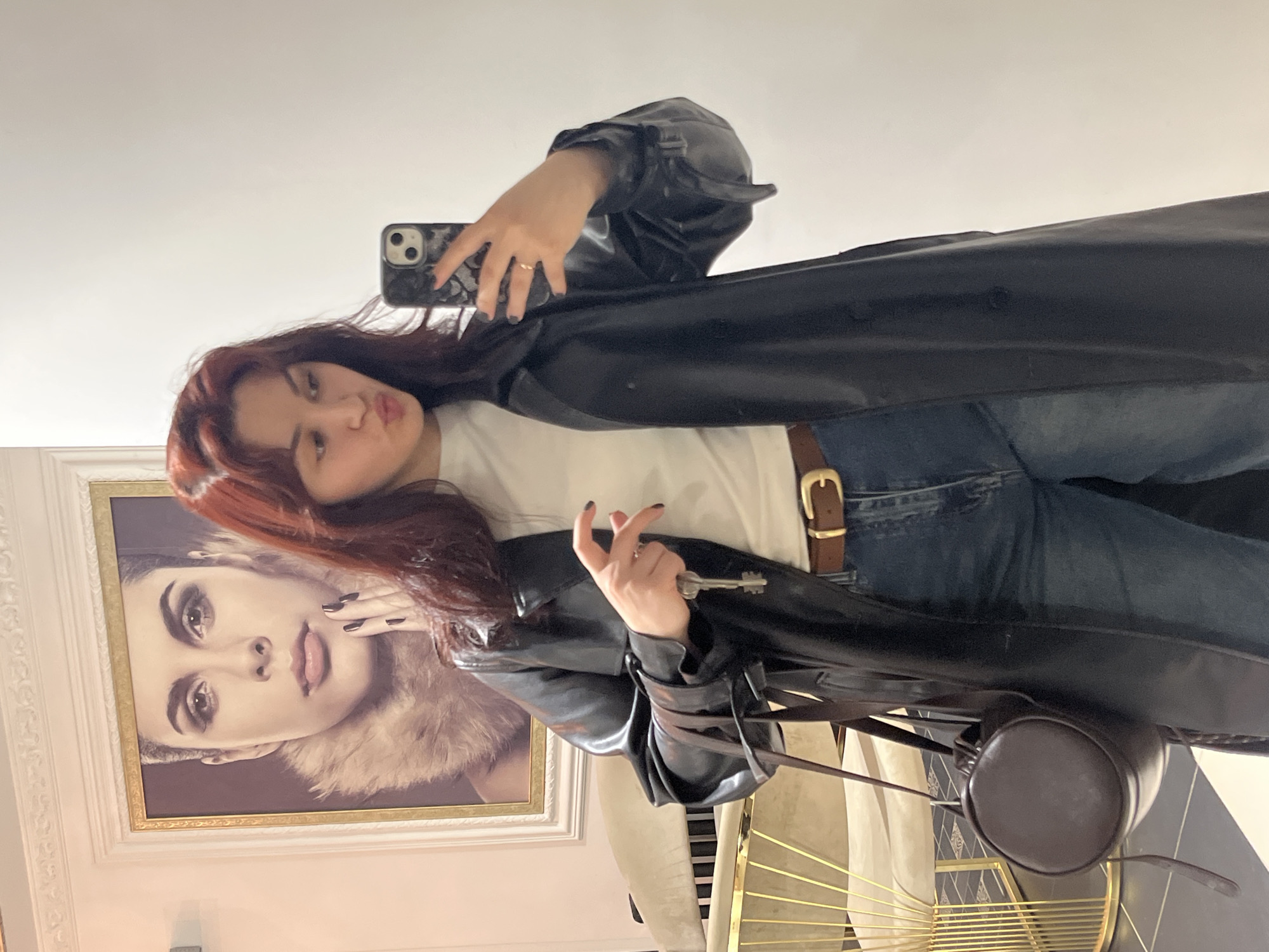 A young woman takes a selfie in front of a mirror. She has red hair and is wearing a black leather trench coat, a white t-shirt, and blue jeans with a brown leather belt. She's holding a key in one hand and a phone in the other. A brown, round purse hangs from her shoulder. Behind her is a framed portrait of a woman with heavy eye makeup and a fur collar. A golden chair is partially visible in the background. The location is likely in Neftekamsk, Russia.