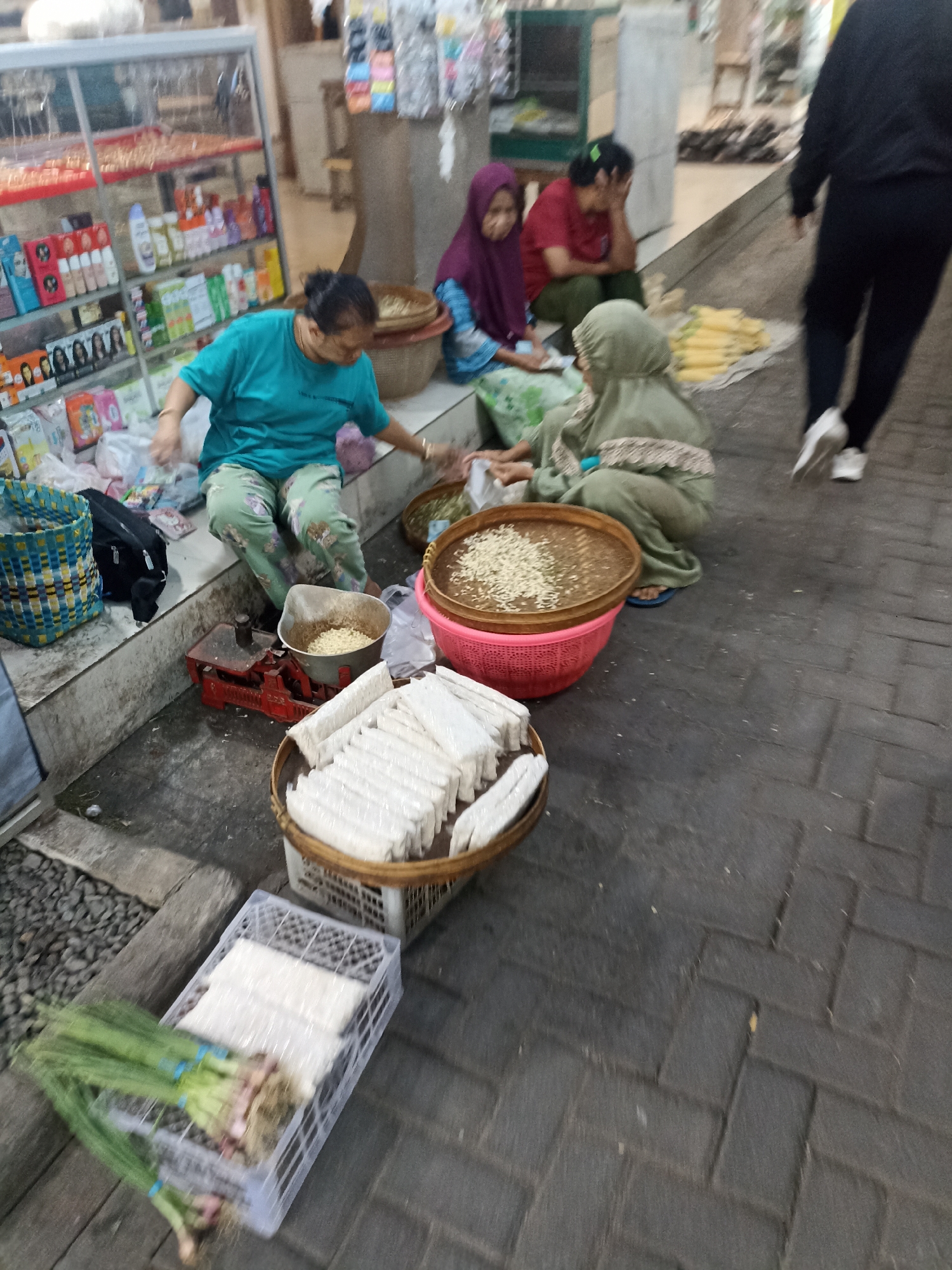 The image depicts a street market scene in Kelurahan Kepatihan, Indonesia. In the foreground, there is a white plastic crate filled with fresh green onions and what appears to be blocks of tempeh wrapped in plastic. Adjacent to this is a woven basket holding more blocks of tempeh. To the left, a red mechanical scale is visible, with a metal bowl on top containing a granular substance, likely rice or grains. Behind this, a woman wearing a teal shirt and patterned pants is seated on a raised concrete ledge, reaching towards the scale. Next to her is a black bag. Further back, three women are seated on the ground. One woman in the center wears a purple hijab and a blue and white patterned garment. To her right, another woman in a red shirt and green pants is seated, with her hand on her forehead. To the right of the woman in the teal shirt, another woman in a light green hijab is seated, tending to a large round woven tray filled with a light-colored granular substance. This tray is placed inside a pink plastic basket. In the background, shelves of various products, possibly toiletries or food items, are displayed inside a glass-fronted cabinet. The ground is paved with dark rectangular stones. A person wearing dark clothing and white sneakers is walking away from the scene on the right side of the image. The overall environment suggests a bustling local market.