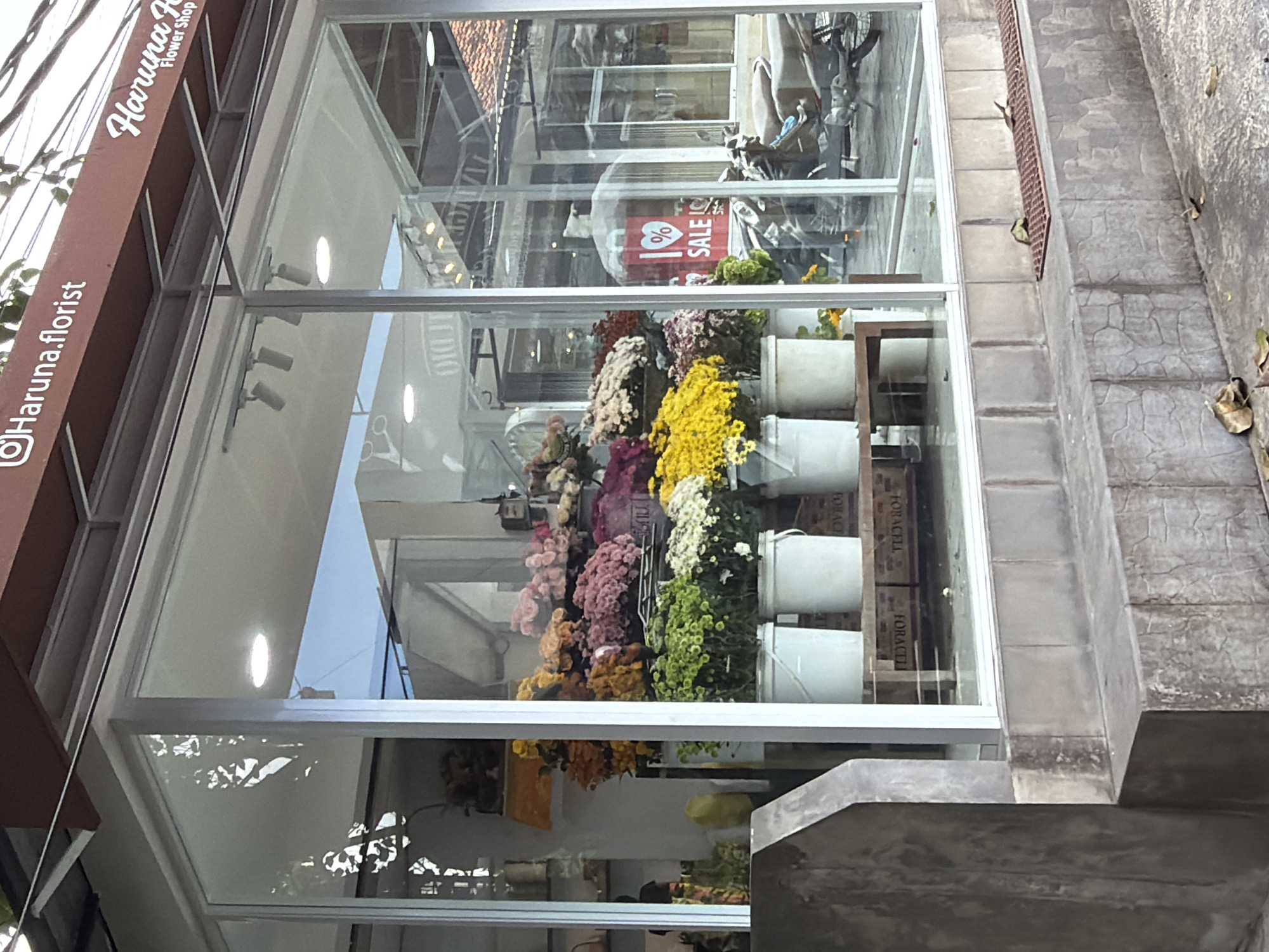 The image shows the storefront of a flower shop called "Haruna Florist," as indicated by the sign on the brown awning visible in the upper left corner. The sign also has "Flower Shop" written on it, along with the Instagram handle "@Haruna.florist."
The shopfront is predominantly glass, allowing a clear view of the interior. Inside, there are rows of flowers in various colors arranged in buckets. A "SALE" sign is visible in the reflection on the glass.
The shop is situated on a street with a gray concrete curb and stairs leading up to the entrance. The reflection in the glass suggests the presence of other buildings across the street.
Based on the information available online, “Haruna florist” is located in North Kuta, Bali, Indonesia.
