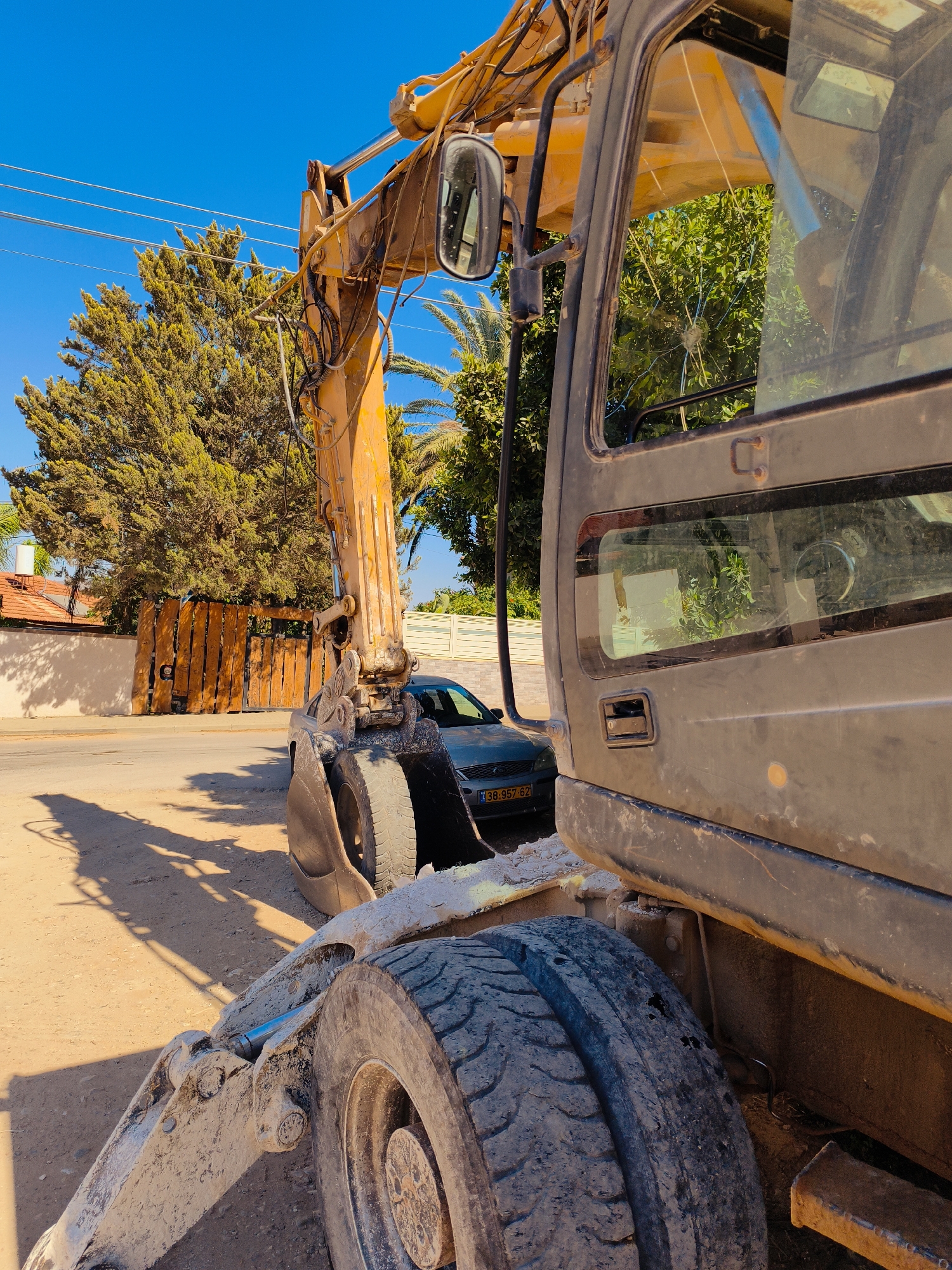 The image shows an excavator parked on a dirt road next to a street. The excavator is yellow and black, with its arm extended. A dark-colored car with an Israeli license plate is parked behind it. In the background, there is a wooden fence and a wall, and behind that, a house with a red tile roof. The sky is blue and clear. The scene appears to be in Lachish Regional Council, Israel, given the style of the license plate and the general environment.