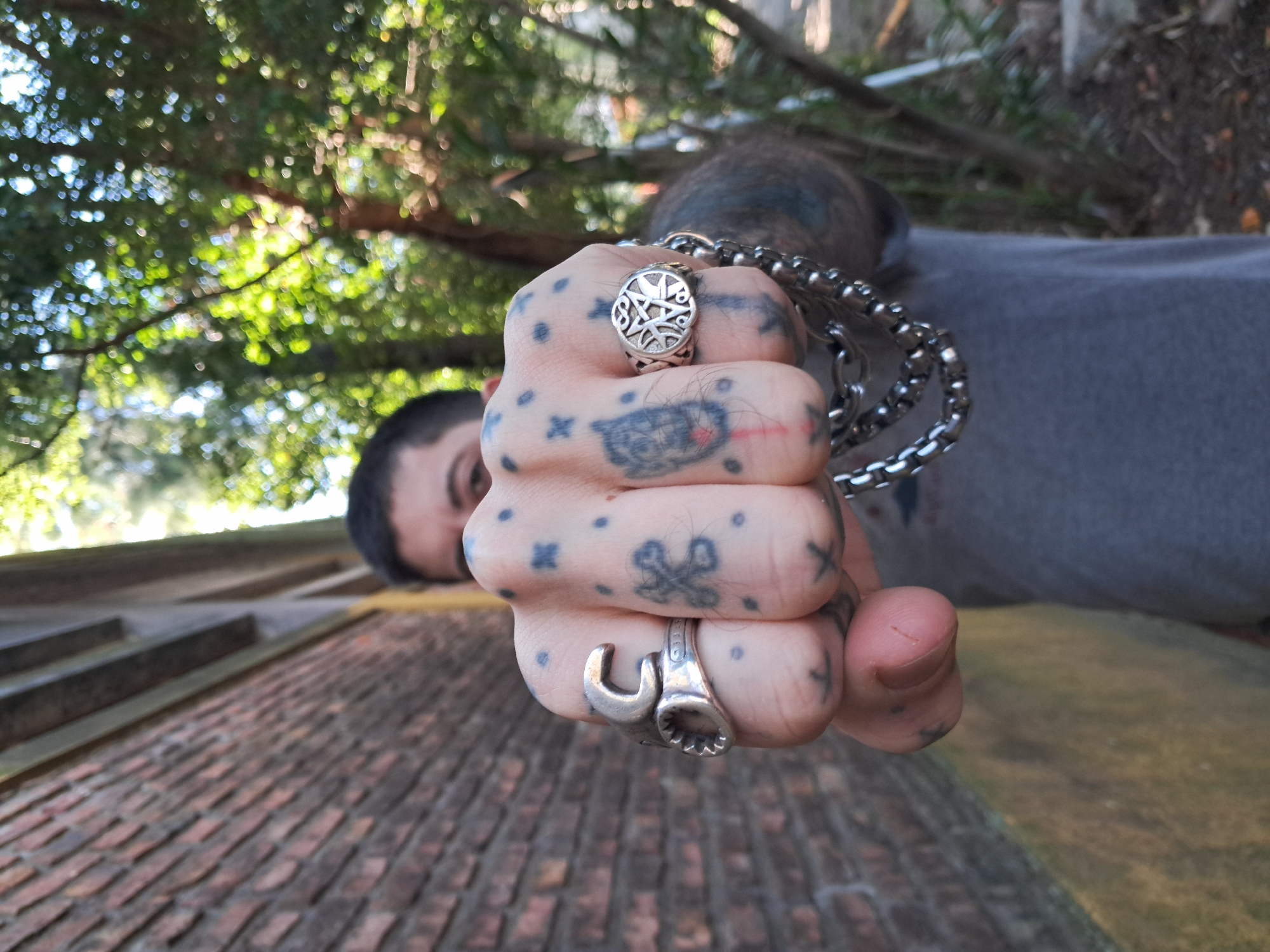 A low-angle, eye-level photograph depicts a clenched, tattooed fist extending towards the camera. The fist is adorned with two rings, one featuring a nautical star design and the other fashioned to resemble two wrenches. A metal chain bracelet drapes over the hand. The person is light-skinned with dark hair.
In the background, visible above the fist, is a portion of the person's face, including an eye, and tattoos visible on the forehead. Further in the background, a brick structure is seen, a patch of greenery appears overhead. The location is Vicente López, Argentina.