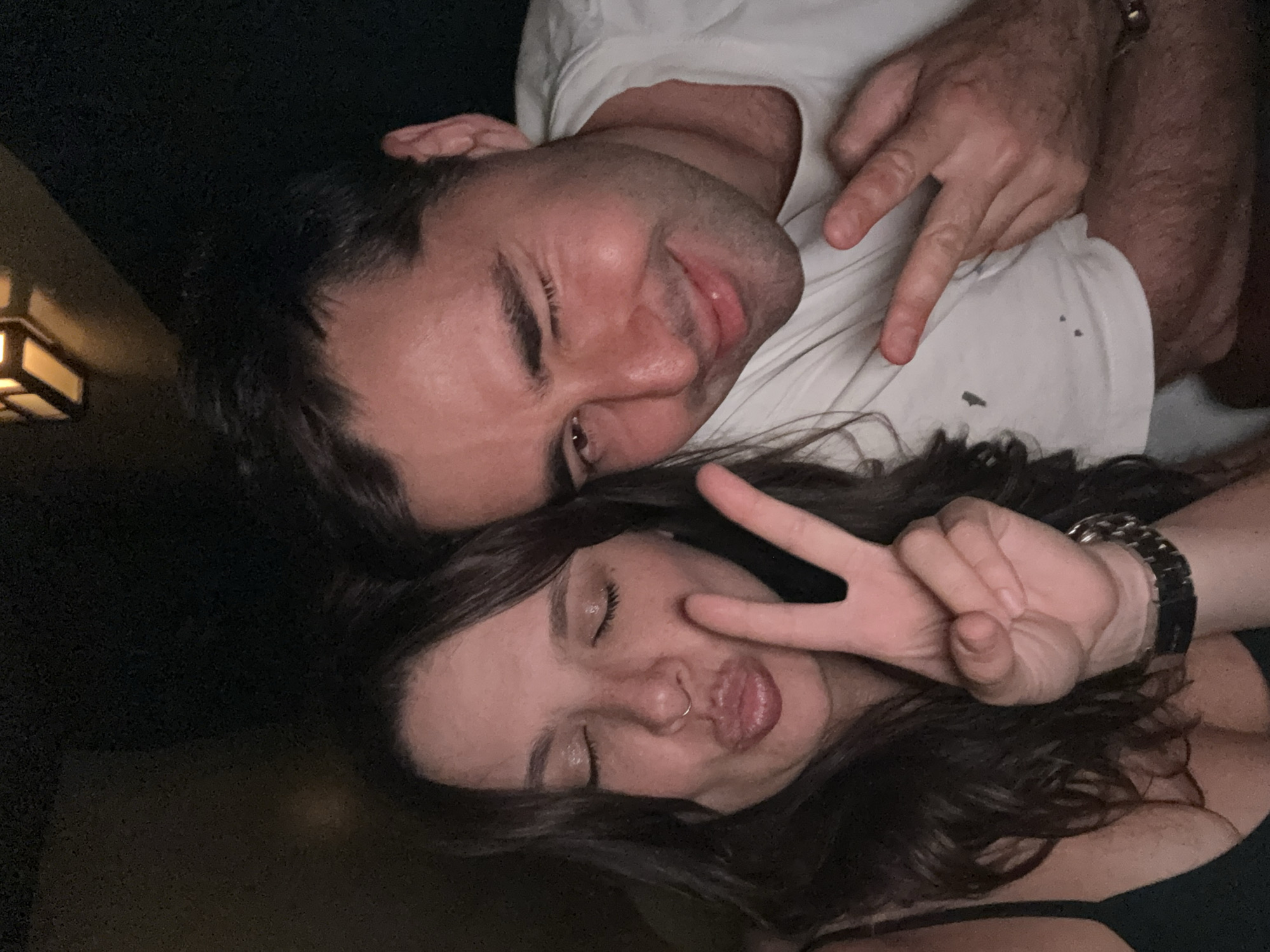Two people are lying side-by-side, appearing to be in an intimate setting, possibly a bedroom or living room. The man is on top, wearing a white t-shirt, and is making a peace sign with his fingers. He has dark hair and appears to be winking. The woman below him has long dark hair and is also making a peace sign, with her eyes closed and puckered lips. A watch is visible on her wrist. The background is dark, with a glimpse of a light fixture in the upper left corner. There is no information in the image to determine the location in Tel-Aviv, Israel.