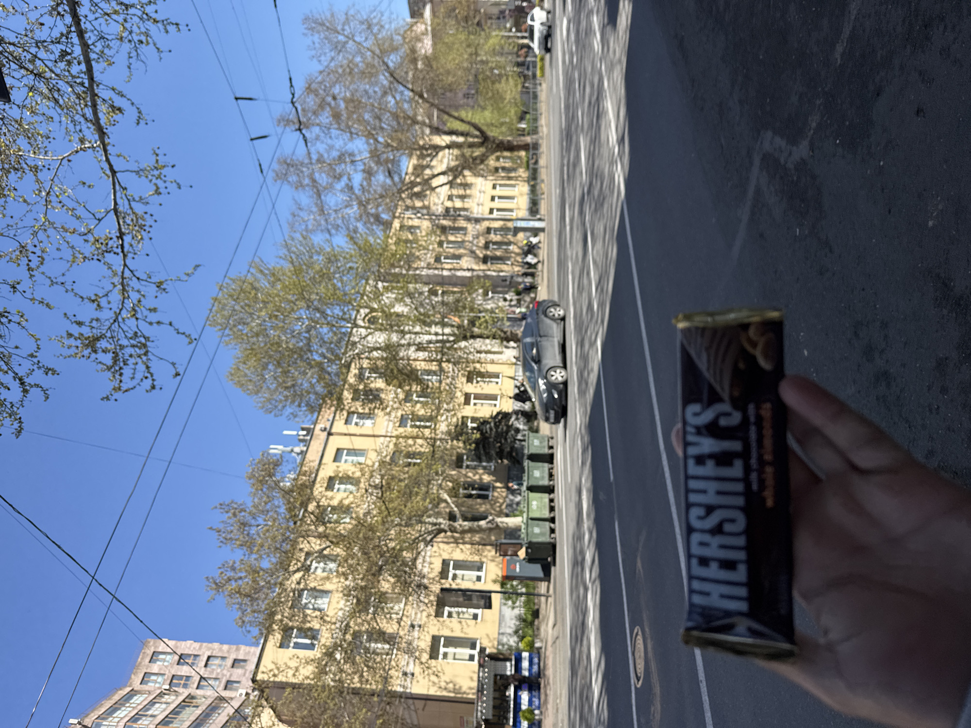 The photograph shows a Hershey's chocolate bar with almonds held in a person's hand against an urban background. The scene is set in Yerevan, Armenia, as evidenced by the architecture and overall urban landscape. The road is asphalt with a white line dividing the lanes. There is a large, multi-story yellow building with many windows. Trees line the street, and there are power lines overhead. Cars are parked on the street. In the distance, there is another building. There is at least one other person present.