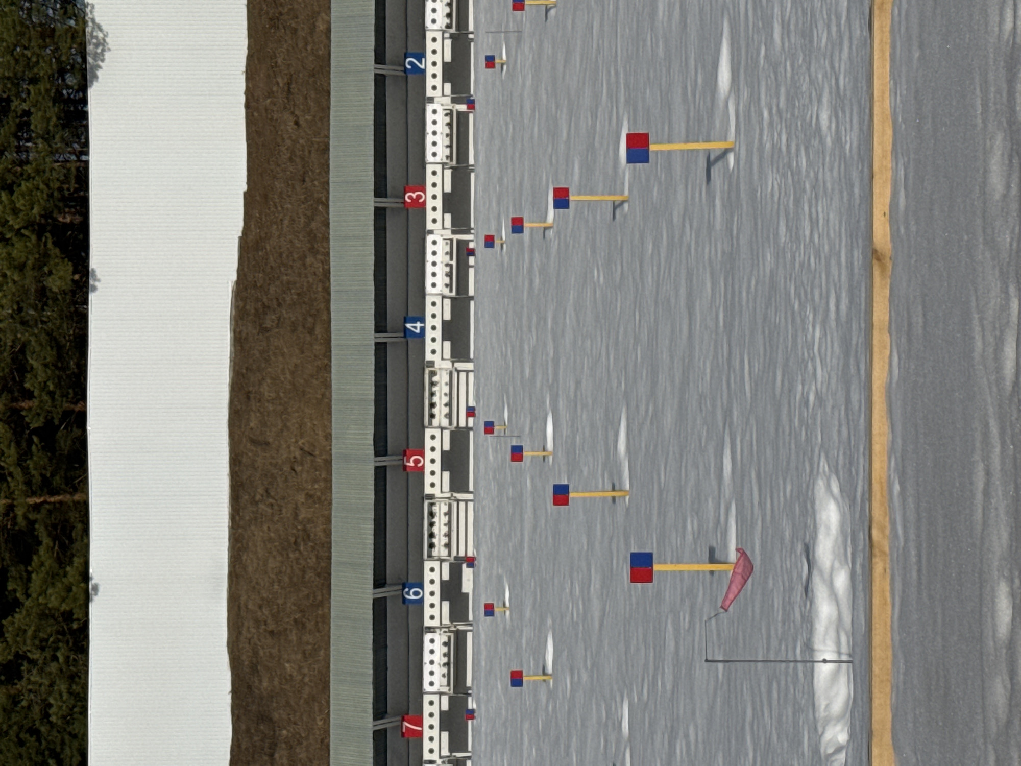 This image presents a forensic view of an empty outdoor biathlon or shooting range in Прикамский, Russia, under winter conditions. The foreground consists of a snow-covered field exhibiting varied textures and subtle undulations. A light brown wooden plank or barrier extends horizontally across the bottom edge of the visible snow. Multiple yellow poles, each topped with a small, rectangular red and blue flag, are arranged in parallel rows across the snow, receding towards the target line. In the lower-left section, a single red windsock hangs downwards from a thin dark pole, indicating minimal wind.
In the mid-ground, a long grey structure with an overhanging roof serves as the target area. This structure contains multiple individual shooting stations, each featuring a white target board with numerous black circular targets. Above these stations, numbered markers are visible: '7' (red), '6' (blue), '5' (red), '4' (blue), '3' (red), and '2' (blue), arranged sequentially from left to right. Behind the target structure, a brown, earthen embankment functions as a backstop. Above this, a tall white corrugated metal barrier provides additional containment. The uppermost portion of the image reveals a dense line of dark green conifer trees against a clear sky. The scene is devoid of any individuals, equipment (other than fixed range elements), or ongoing activities.