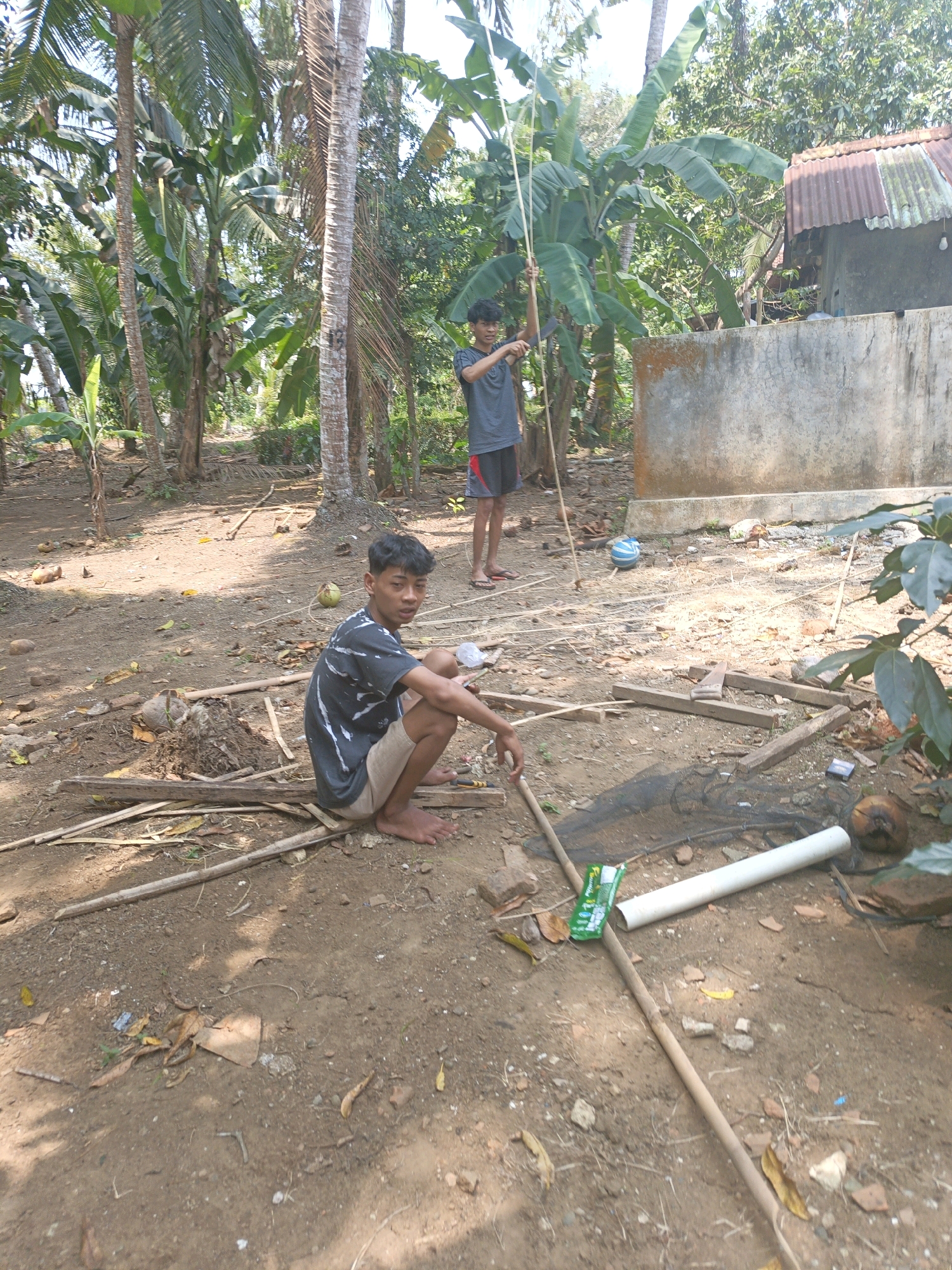 In the outdoor setting of Kamulyan, Indonesia, two young boys are engaged in an activity involving bamboo poles. One boy, wearing a dark t-shirt with a graphic print and light-colored shorts, is seated on the ground, barefoot. He is positioned amidst scattered bamboo poles, wood scraps, and dried leaves. To his right, a white PVC pipe and a green packet of what appears to be crackers are visible on the ground, near a black net. Another boy, dressed in a gray t-shirt and dark shorts with red trim, stands further back. He is holding a long bamboo pole upright, seemingly in the process of using it. The environment is rustic, characterized by dirt ground, numerous bamboo and palm trees, and a simple concrete structure with a corrugated metal roof in the background. Coconuts are scattered on the ground, suggesting a tropical locale.