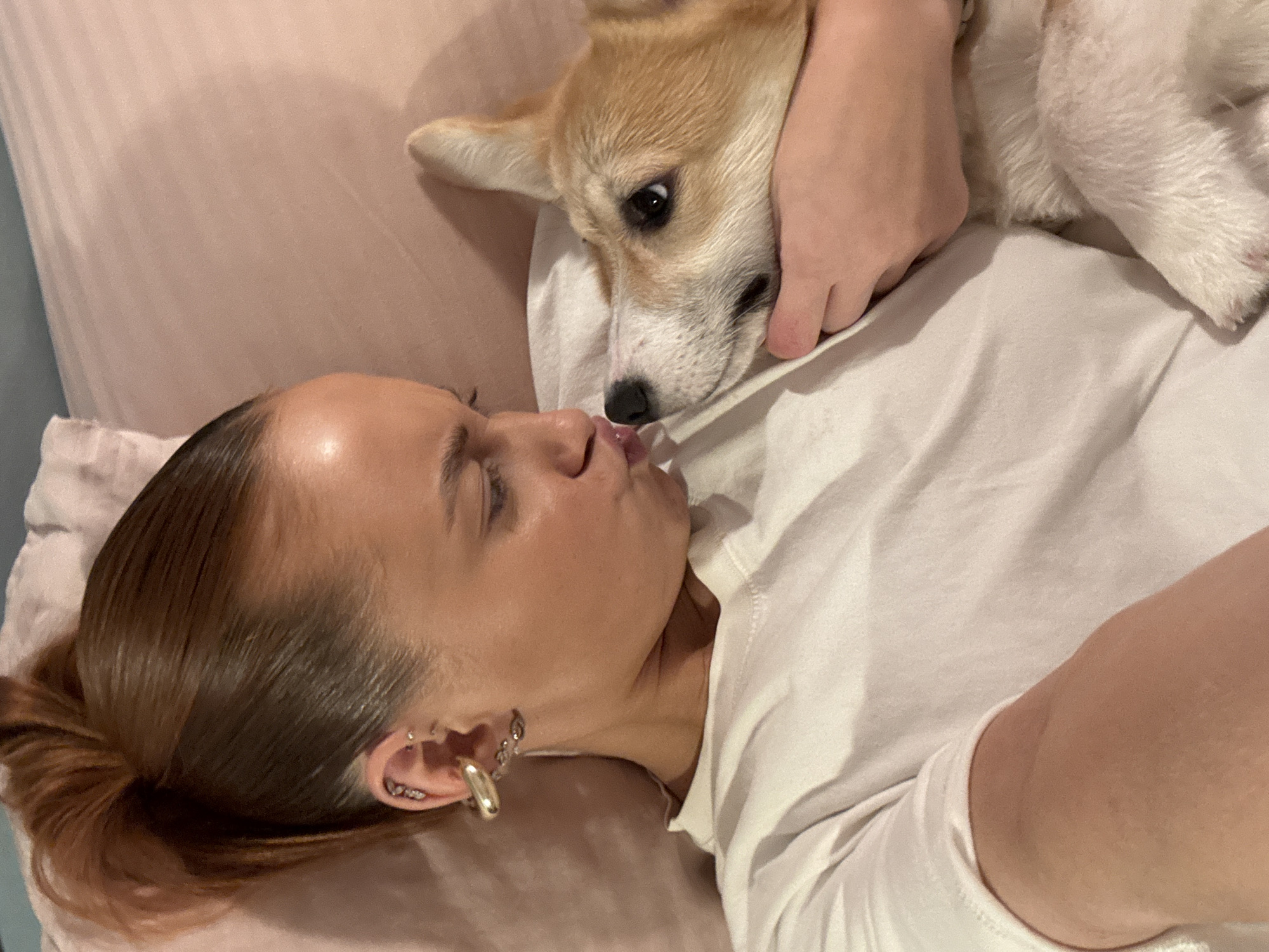 A woman with brown hair in a low ponytail and multiple ear piercings is lying on a pink bed, wearing a white t-shirt. She is looking up at and puckering her lips towards a corgi puppy being held above her by an unseen person. The location is unknown, but the prompt suggests it may be Chaikovsky, Russia.