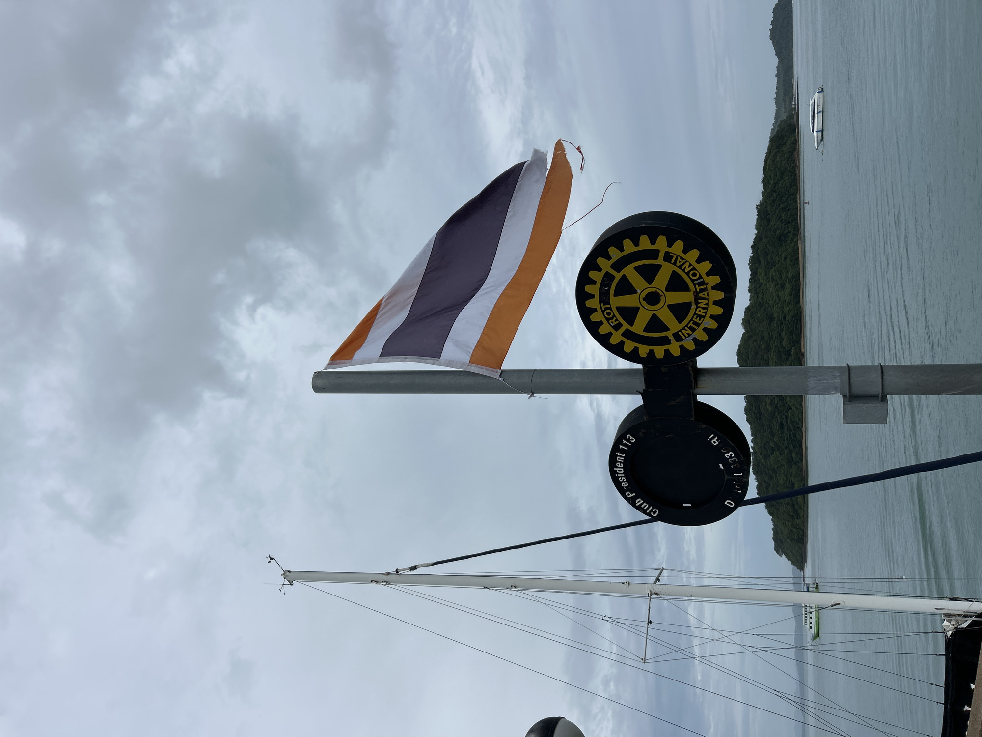 In Pa Khlok, Thailand, an overcast sky looms above a serene bay. The foreground is dominated by a flagpole flying the Thai flag, its orange and white stripes catching the muted light. Below the flag, a circular sign with a Rotary International emblem is mounted on a pole, flanked by another sign with text that appears to read "Club President 113" and "District 3330 Ri". To the left, the mast of a sailboat stands tall, its rigging a complex web against the cloudy backdrop. In the distance, lush green islands dot the horizon. A white speedboat is anchored in the tranquil waters, and a larger green and white ferry navigates the bay further out. The scene captures a moment of quiet maritime activity under a diffused sky.