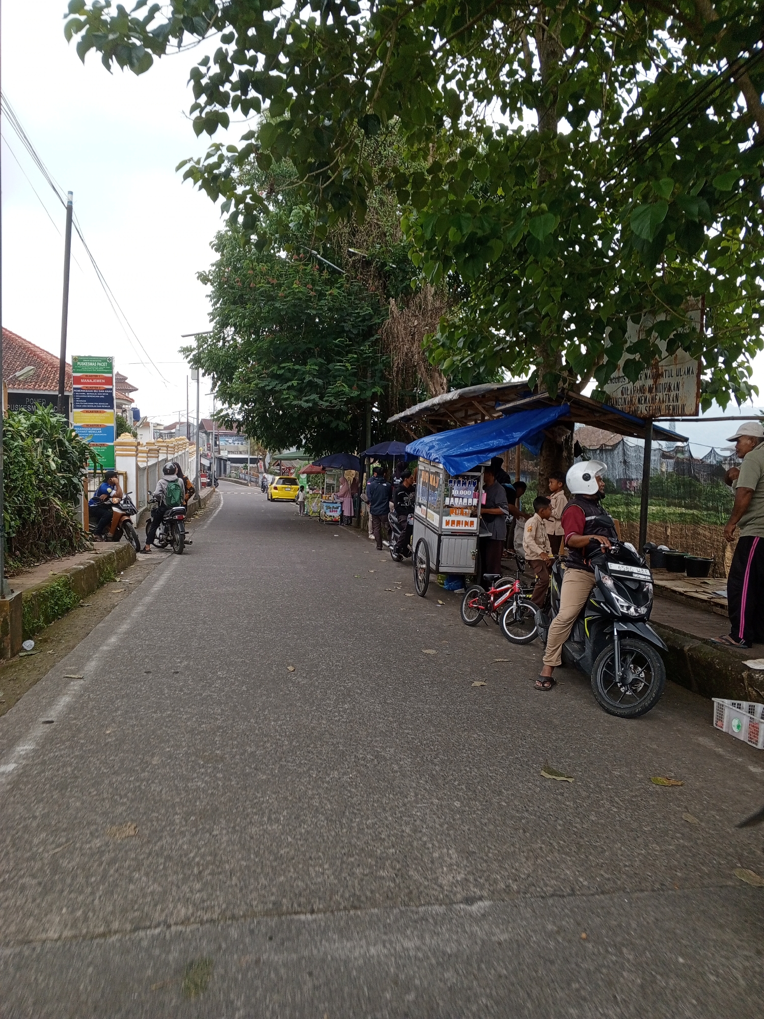 The image depicts a lively street scene, likely during daytime under an overcast sky. The main subject is a moderately busy asphalt road that gently curves to the left, lined with various activities and structures. On the right side of the road, several street vendors are operating under makeshift stalls, identifiable by their blue tarpaulin roofs. One vendor's cart, likely selling food or drinks, is prominently visible, and a person in a white helmet is seated on a black motorcycle parked beside it. A small red child's bicycle is also near the cart. Several individuals, including children in school uniforms, are gathered around these stalls, indicating commerce.
Further down the street on the left, two men are sitting on motorcycles near a low wall. The area is densely vegetated with large green trees providing shade, and overhead power lines are visible. Residential or commercial buildings with red-tiled roofs are seen in the background on the left.
Visible text on various signs includes "PUSKESMAS PACET," "KLASTER," "MANAJEMEN," "ANAX," "ANGAN," "10.000," and "KERINE." Another sign partially reads "ENGURUS... NATATUL ULAMA DILARANG MENDIRIKAN BANGUNAN MEMANFAATKAN TANPA SEHIN." A motorcycle license plate clearly displays "F 6580 HAE." The scene suggests a common daily life in a community, with local businesses thriving by the roadside.