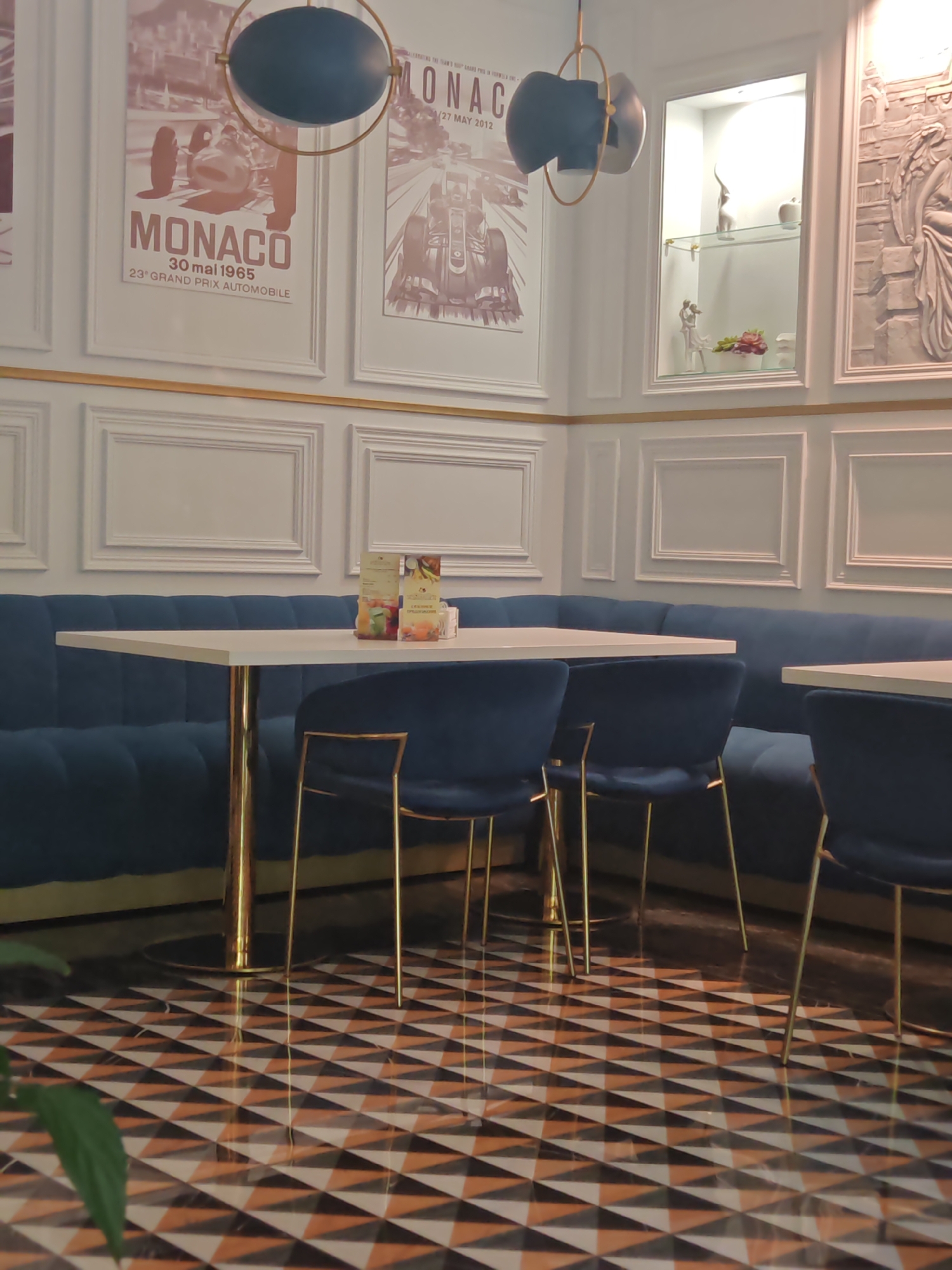 This image captures the interior of an elegant cafe or restaurant in Sochi, Russia, featuring a modern classic design. The scene is devoid of people, suggesting it might be unoccupied.
A plush, deep blue velvet booth seating area lines the walls, complemented by white tabletops with golden pedestal bases and matching blue velvet chairs with slender gold legs. The walls are adorned with white panel molding and a thin golden trim running horizontally. Several framed posters, rendered in a sepia tone, depict vintage and modern Monaco Grand Prix automobile races. One poster clearly reads "MONACO," "30 mai 1965," and "23e GRAND PRIX AUTOMOBILE." Another poster nearby also mentions "MONACO" and "27 MAY 2012."
In the corner, a recessed white display cabinet with glass shelves showcases decorative items, including small white figurines and a floral arrangement, illuminated by internal lighting. Above, a contemporary light fixture with blue and gray elements and a golden frame hangs from the ceiling.
The floor is a striking feature, covered in geometric patterned tiles composed of alternating black, white, and brown triangles, creating a dynamic visual effect. A green plant is partially visible in the foreground. Menus and small promotional items are placed on one of the tables. The overall ambiance suggests a sophisticated and well-maintained dining space.