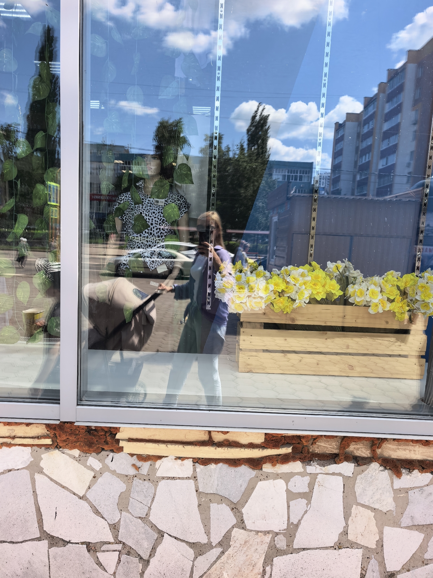 The image shows a reflection in a storefront window. Inside the store, a mannequin is displayed wearing a black and white leopard print top. A woman is visible in the reflection, holding a stroller and taking a photo of herself with her phone. In front of the window, there is a wooden planter box filled with yellow and white flowers. The storefront is located in the city of Sterlitamak, Russia. The exterior of the building features a wall made of large, irregularly shaped stones and bricks. The sky is blue with white clouds, and a building is visible in the background.