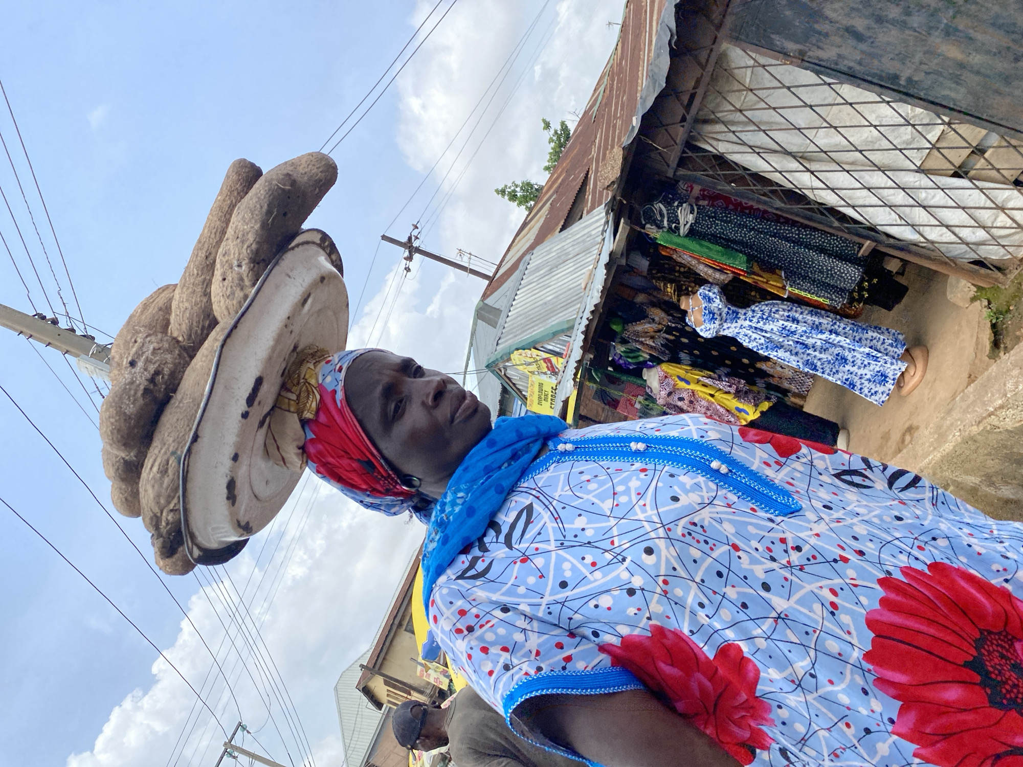 An outdoor market scene in Bukuru, Nigeria, features a woman balancing a large quantity of yams on her head, supported by a white tray. She is dressed in a light blue, patterned tunic with red floral accents and a blue scarf. To her right, a stall displays various colorful dresses. In the background, other market stalls and a few people can be seen. Overhead, power lines crisscross the sky, and corrugated metal roofs are visible. The overall setting suggests a typical bustling Nigerian marketplace under a cloudy sky.
