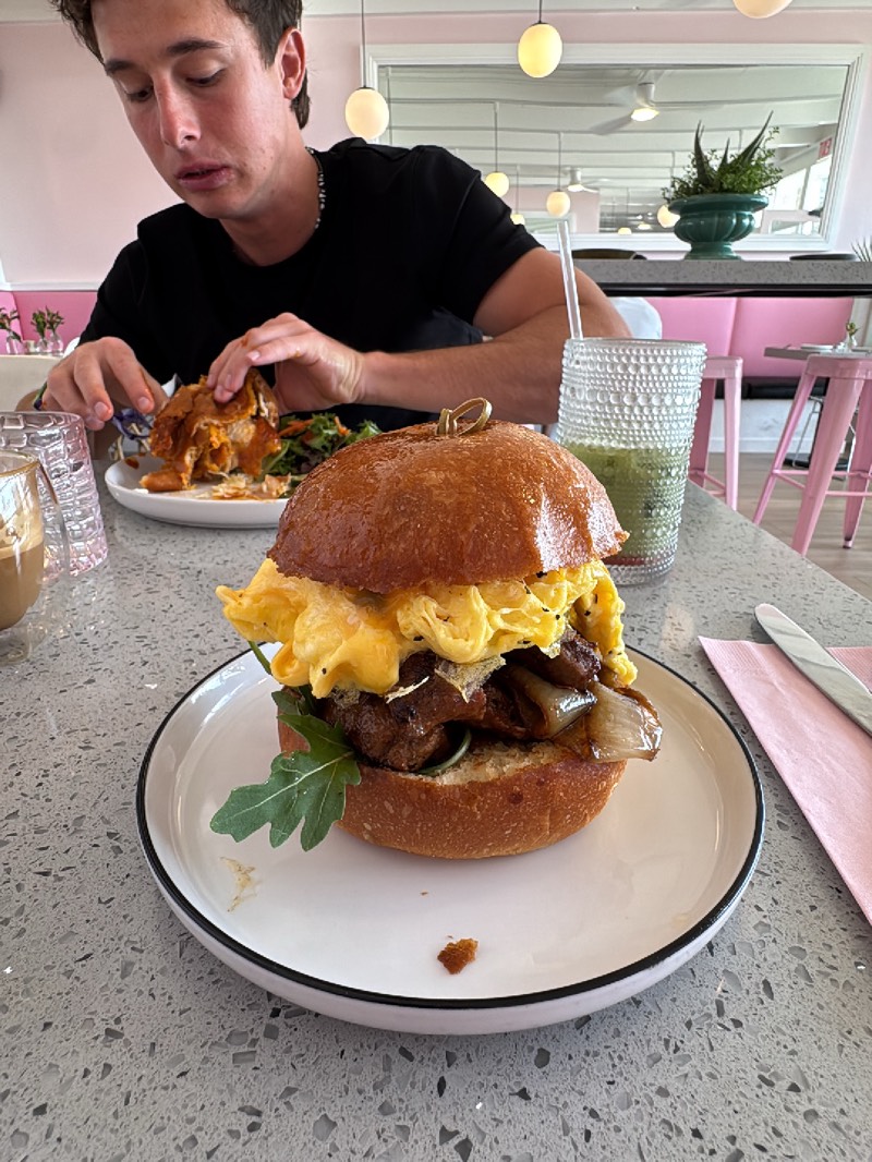 The image shows a man eating a meal at a restaurant in Palm Desert, California. The man is white, has short brown hair, and is wearing a black t-shirt. He is eating a dish with his hands, and there is a plate with salad next to it. In the foreground is a hamburger on a white plate. The hamburger is topped with scrambled eggs, grilled onions, and arugula. Next to the burger, there is a pink napkin and a knife. In the background, there is a pink booth and a table with a succulent plant. The restaurant has pink and white decor.