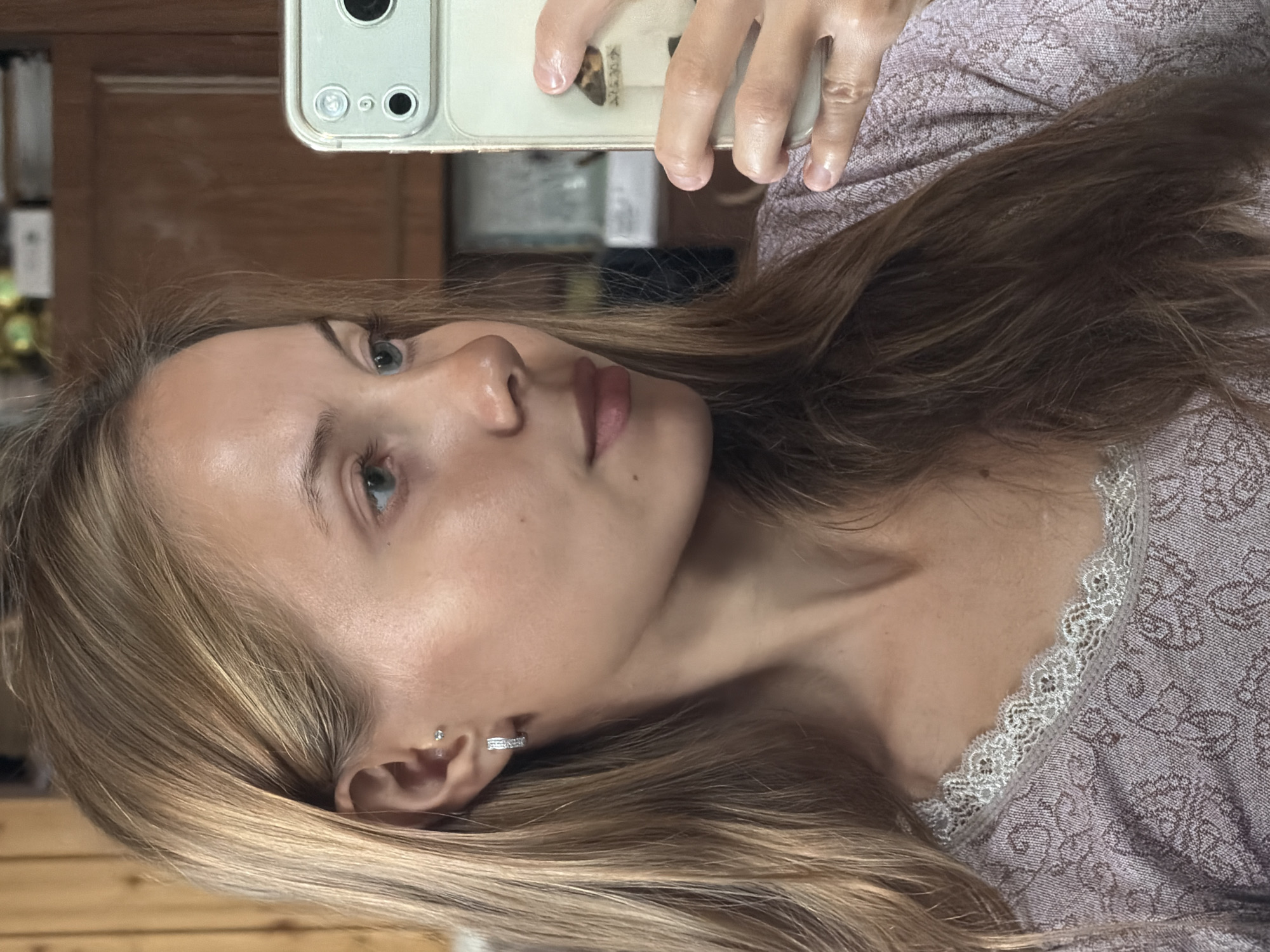 A young woman with blonde hair and blue eyes is taking a selfie in a room with wooden walls. She is wearing a purple top with lace trim. She has two piercings in her left ear, one with a small stud and another with a hoop earring. In her right hand, she is holding a smartphone with a light green case. A small sticker of a moth is attached to the back of the phone case. The lighting in the room appears to be natural and is coming from the left side of the frame.