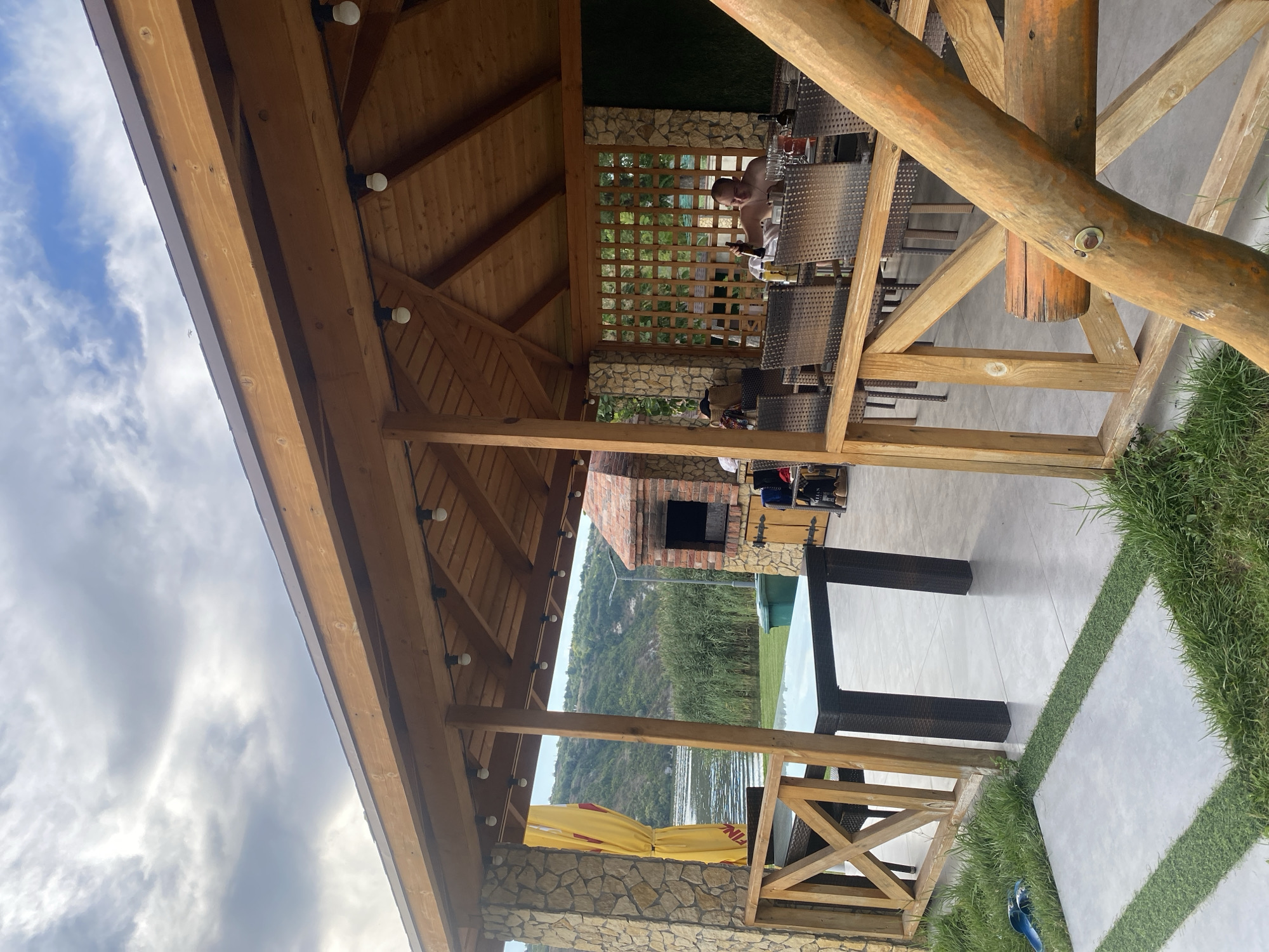 This is a medium shot photo of an outdoor dining area at a restaurant or resort in Molovata Nouă, Moldova. The photo is taken from a low angle, looking up towards the wooden roof of the structure.
In the background, a river and hills covered in trees can be seen. A brick barbecue pit is visible to the left of the dining area, and there is a picnic table with chairs to the right. A man is seated at the table, holding a bottle of beer. There are also some bottles and glasses on the table. The sky is cloudy.