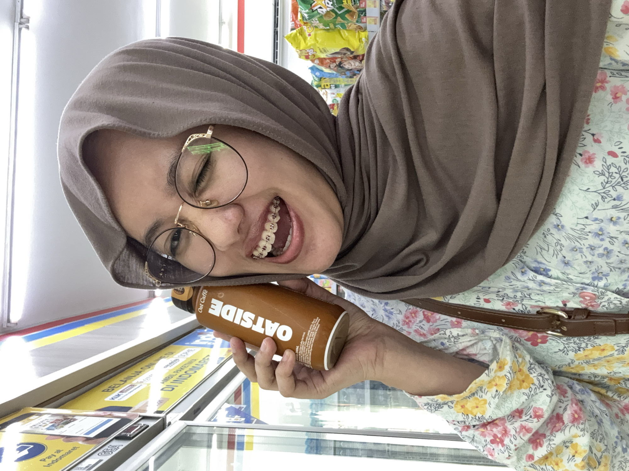 A young Indonesian woman, wearing a brown hijab and a floral patterned shirt, is taking a selfie in what appears to be a convenience store in Gunungsari, Indonesia. She is holding a bottle of "OATSIDE" oat milk drink up to her ear, smiling with her mouth open, revealing braces. The background shows shelves stocked with various products, including bags of snacks and advertisements for services like "Pay at Indomaret." The lighting suggests an indoor, well-lit environment typical of a retail establishment.