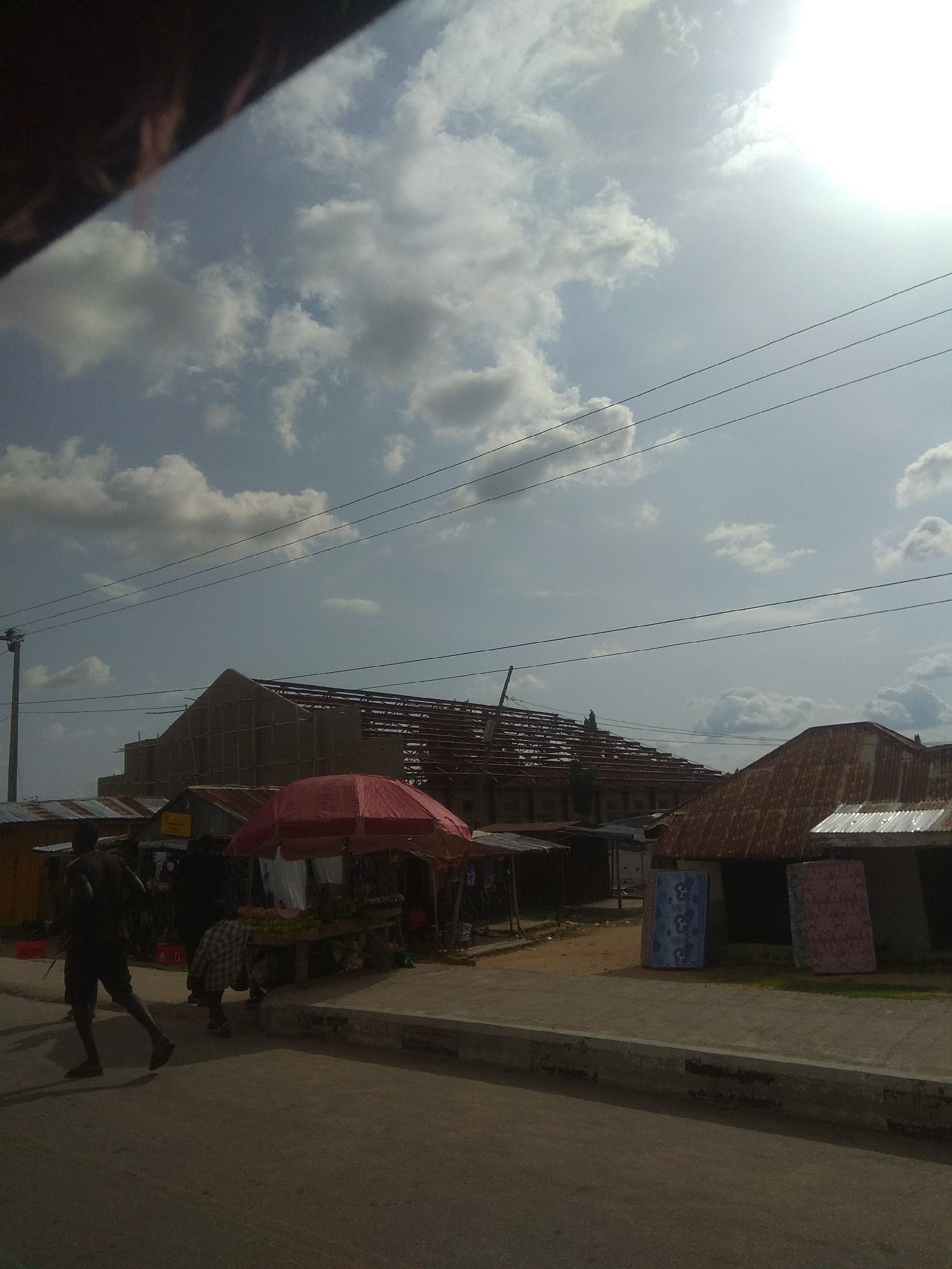 The image depicts a street scene in Jalingo, Nigeria, under a bright, partly cloudy sky, suggesting it is daytime. A large building under construction, with its roof framework exposed and scaffolding around it, dominates the background. Several smaller structures with corrugated iron roofs are situated in front of it, some appearing to be market stalls.
In the foreground, a person is walking or running on the road, their silhouette dark against the pavement. To the left, near the market stalls, another person is bent over, possibly engaged in activity related to the market. A red umbrella shades a stall where produce, possibly fruits, are displayed on a table. Two mattresses, one blue and one pink with patterned covers, are propped up against a building on the right side of the image.
Power lines are visible crisscrossing the sky, and a utility pole is present on the far left. The overall impression is one of a bustling, possibly commercial area with ongoing construction and daily commerce. No discernible text is visible in the image.