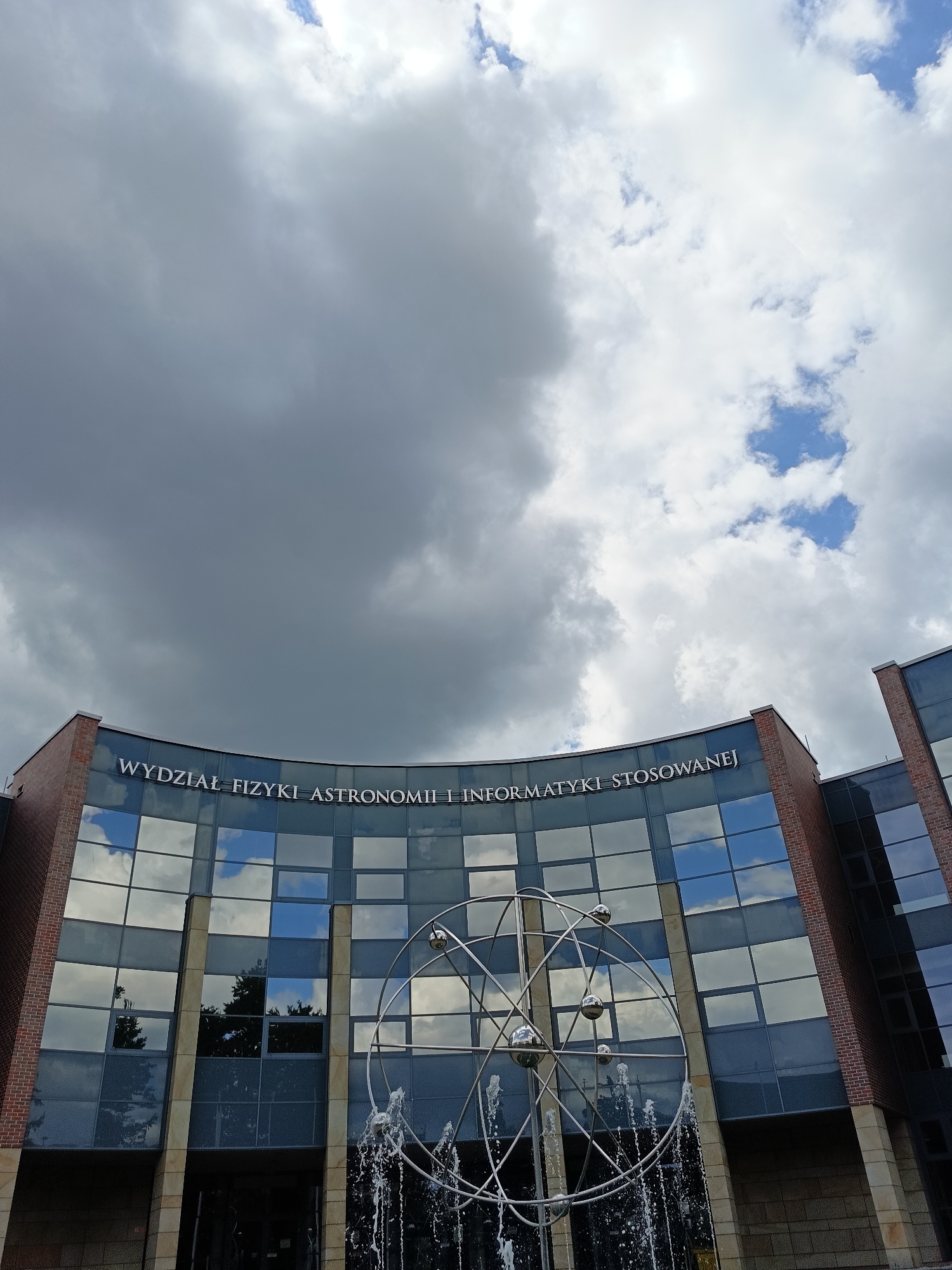 The image captures the exterior of a modern building with a curved glass facade, likely an academic institution, set against a dramatic sky filled with a mix of dark and light clouds. White lettering across the top of the glass reads "WYDZIAŁ FIZYKI ASTRONOMII I INFORMATYKI STOSOWANEJ," which translates to "Faculty of Physics, Astronomy and Applied Informatics." In front of the building, a metallic sculpture resembling an atomic structure, with spherical elements and radiating lines, is partially obscured by a fountain with cascading water. The building's brick and stone exterior is visible on the sides. The overall impression is one of a contemporary scientific or educational facility under a dynamic sky. Based on the text on the building, this location is the Faculty of Physics, Astronomy and Applied Informatics at Jagiellonian University in Krakow, Poland.