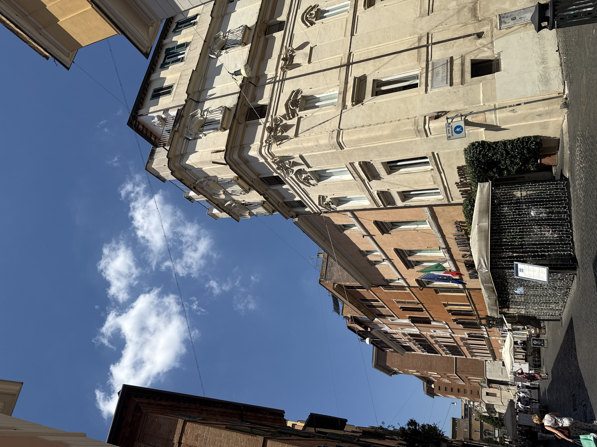 This is a street-level, medium-angle, full shot of a narrow street in Rome, Italy. The street is paved with cobblestones and lined with tall, old buildings. The sky is a clear, bright blue with scattered white clouds.
On the right side of the street is a large, ornate, cream-colored building with multiple windows and balconies. The building has decorative architectural elements, including moldings and pilasters. A street sign on the building reads "VIA DEL FALCO".
To the left of this building, the street continues, lined with other buildings of varying heights and colors, predominantly orange and brown brick. Some of these buildings have shops or restaurants at ground level, with awnings and outdoor seating. Flags are visible hanging from one of the buildings.
There are a few people walking on the street, including a woman in the foreground on the left, carrying a bag. Some people are seated at tables outside the establishments. The scene appears to be bustling with activity, likely a tourist area.
In the foreground, a covered outdoor seating area is visible, decorated with string lights. A metal railing encloses the area. A sign is posted on an easel within this enclosure.
The overall impression is a sunny, lively day in a historic European city.