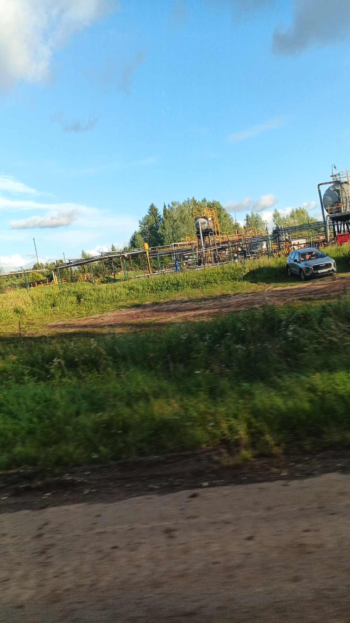 The image depicts an industrial site with extensive piping systems elevated on yellow and green supports. The site appears to be situated in a rural or semi-rural area, with a backdrop of green trees. A silver car is parked on a dirt path adjacent to the industrial infrastructure. The sky is blue with scattered white clouds. The foreground shows a dirt road or path with blurred green grass on either side, indicating the photo was taken from a moving vehicle.
