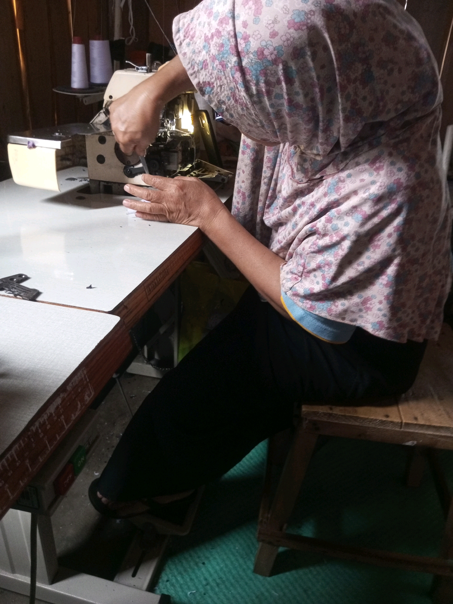 An Indonesian woman, wearing a floral headscarf and a pink floral patterned blouse with a light blue collar, sits on a wooden stool in front of a sewing machine in a workshop in Kalibeber, Indonesia. Her hands are on the sewing machine, and she is operating it. The sewing machine is on a white table with a wooden edge. There are spools of white thread on a shelf behind the sewing machine. The floor is covered with a green rug. The lighting is dim, with some light coming from the sewing machine.