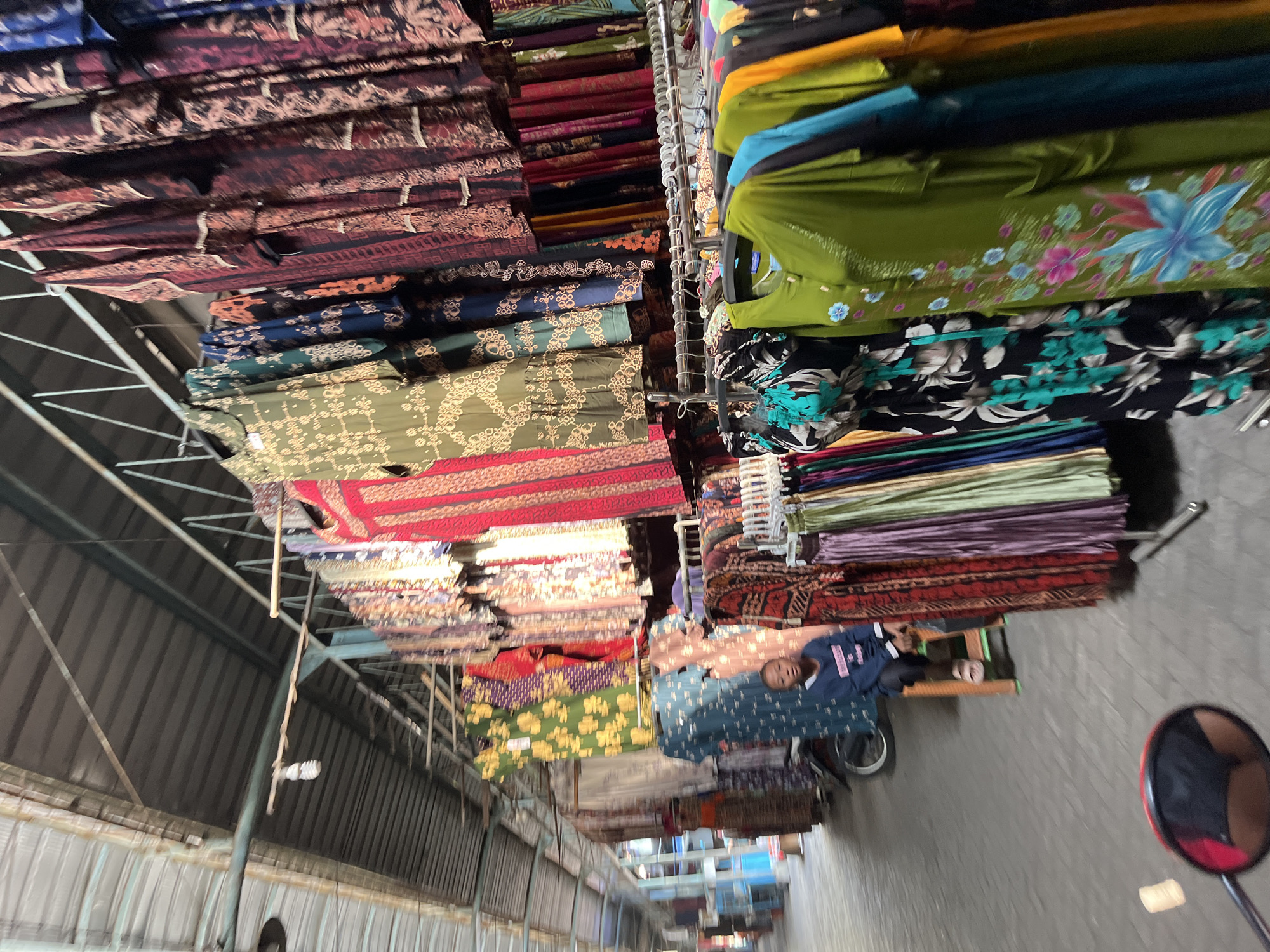 A young person sits on a small stool at a market stall in Juwet Kenongo, Indonesia. The stall displays a variety of colorful, patterned dresses hanging from racks, as well as other garments draped over a motorcycle. The market appears to be covered by a corrugated metal roof, with metal beams supporting the structure. The ground is paved with what looks like stone or concrete. The person, wearing a dark blue sweatshirt and light-colored sandals, looks towards the camera. The image is taken from a low angle, possibly from a motorcycle, as a red rearview mirror is visible in the foreground.