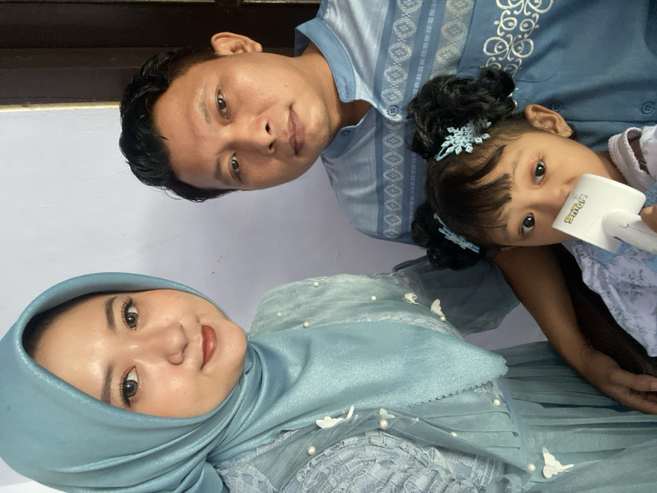 Three individuals, two adults and one child, are depicted in a frontal, chest-up portrait indoors in Tambakrejo, Indonesia, against a plain light-colored wall with a dark brown vertical doorframe visible on the right. The woman on the left wears a light blue hijab and a matching dress with lace overlays, white pearl accents, and white butterfly appliques; she looks directly at the camera with a slight smile. The man on the right wears a light blue collared shirt with darker blue geometric patterns and also looks directly at the camera with a neutral expression. A young girl, positioned centrally below the adults, has dark hair styled in two pigtails with light blue snowflake-shaped hair clips and wears a light-colored patterned top. She holds a white, rectangular object with "S+ Plus" branding in her right hand and looks slightly upward and to the right, away from the camera.