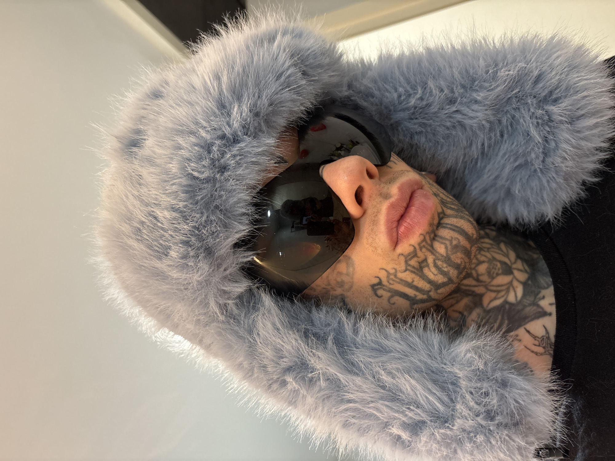 A person wearing a light blue fur hat and dark sunglasses looks directly at the camera from a low angle. The person has many tattoos on their face and neck. The background is a slightly blurred white or light grey. The image does not contain information about the location in the city of Buenos Aires, Argentina.