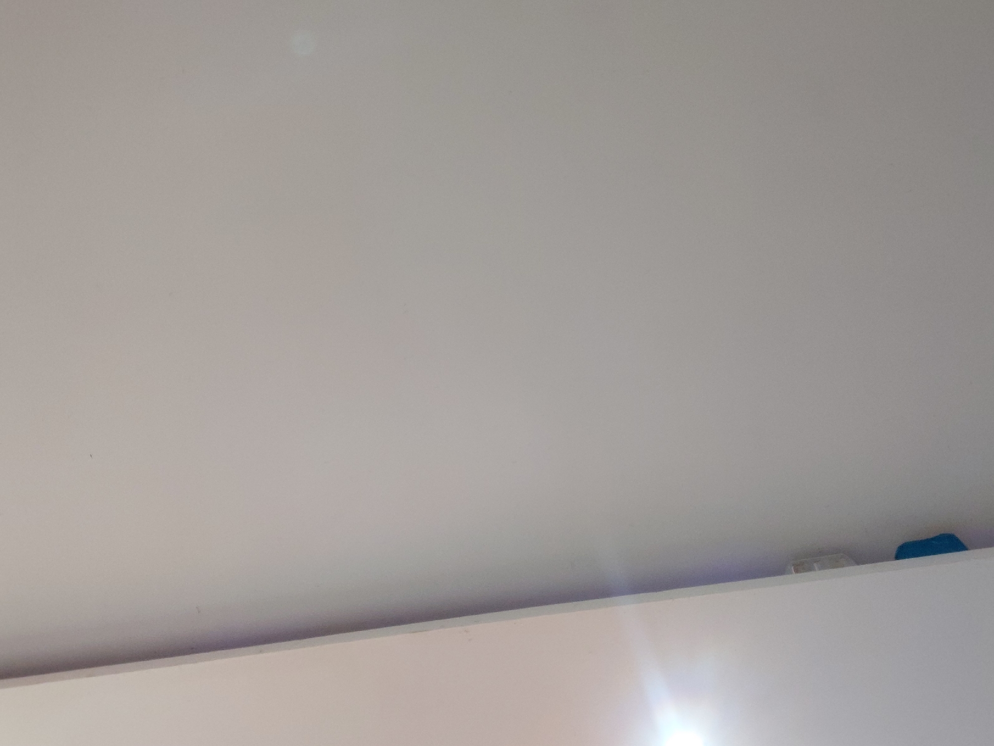 This image captures an upward-facing view of a plain, light-colored ceiling and the upper edge of a wall or shelf in an indoor setting in Santa Ana, Venezuela. The majority of the frame is dominated by the off-white or very light grey ceiling, which appears mostly unadorned. A subtle, hazy light reflection is visible towards the upper left portion of the ceiling.
Along the bottom of the image, a horizontal line marks the transition to a lower surface, which is brightly illuminated. This appears to be the top edge of a wall or a piece of furniture, possibly a high shelf or cupboard. On this narrow surface, near the right side, two small objects are discernible. One is a light-colored, possibly white, rectangular or box-like item. Next to it, to the right, is a distinct blue object with a somewhat irregular shape, possibly a piece of cloth or a small container.
A prominent lens flare or strong glare from a light source, likely out of frame, emanates from the bottom right corner, casting a bright streak across the lower portion of the image. No people are visible, and no specific activity is taking place. The scene is static, depicting a simple interior space without any visible text or distinct architectural details to indicate a specific room type or time of day beyond the presence of light.