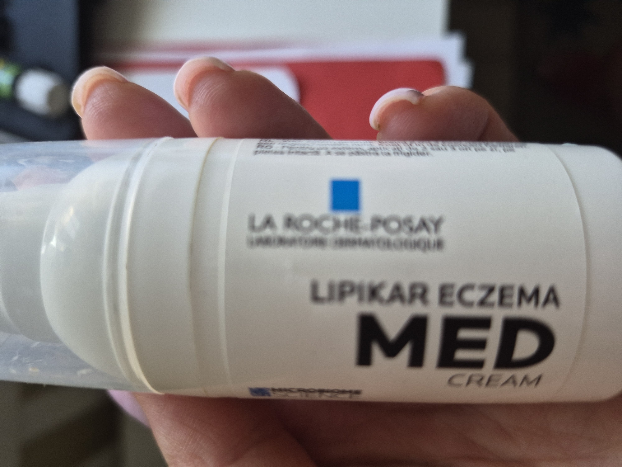 The media file is a close-up shot of a person's fingers holding a white tube of cream. The tube has a clear plastic dispenser and a white label. Visible text on the label includes "LA ROCHE-POSAY," "LABORATOIRE DERMATOLOGIQUE," "LIPIKAR ECZEMA MED," and "CREAM." There is also a blue square logo and a smaller logo with the word "MICROBIOME" visible. The lighting suggests it is daytime. The background is blurred, but appears to be an indoor setting with some red and dark objects. The focus is on the product and the fingers holding it, implying the subject is about to use or has just used the cream.