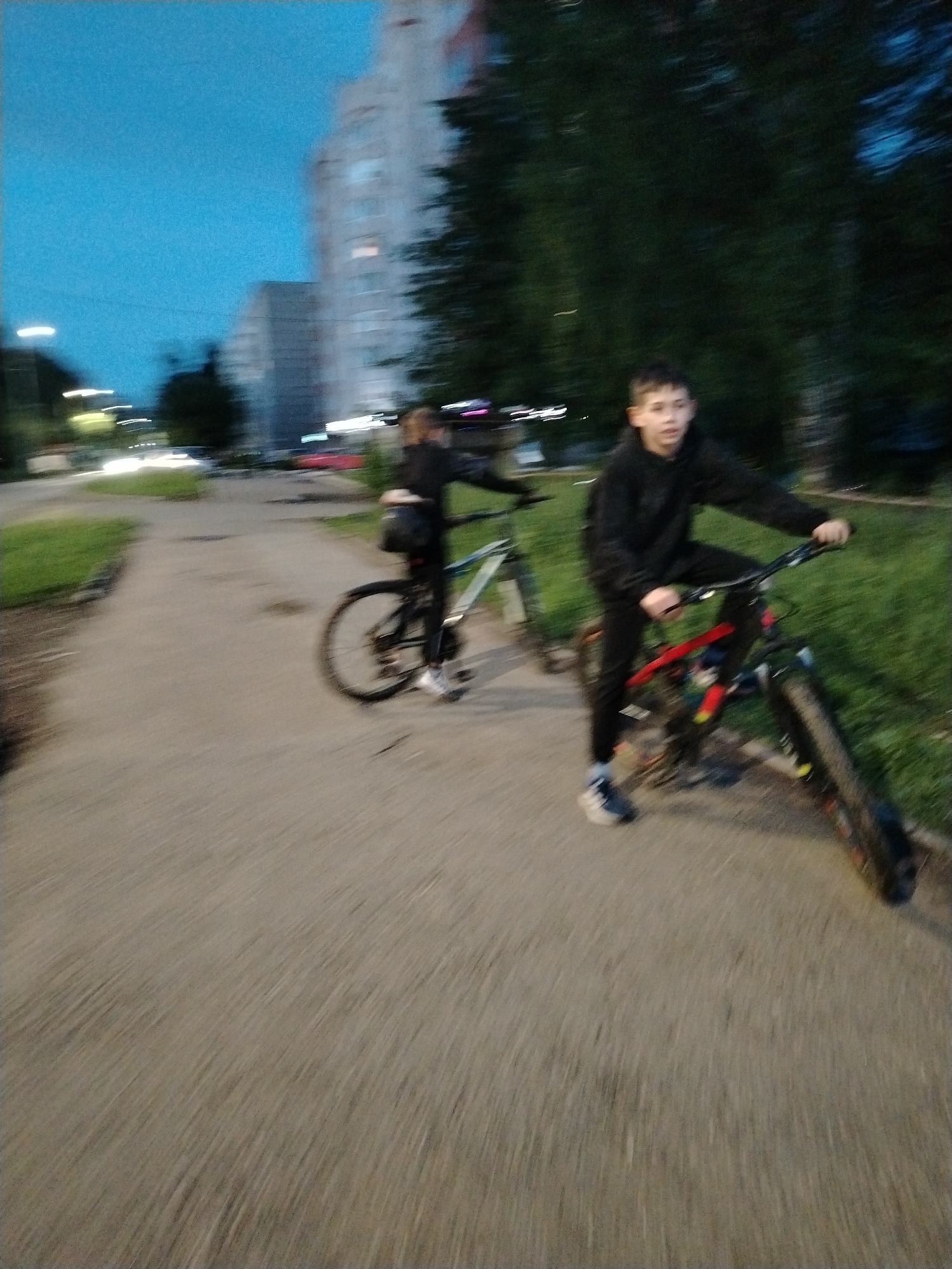 Two individuals are depicted on bicycles in Izhevsk, Russia. The scene occurs along a paved pathway lined with grass on either side. In the background are apartment buildings and trees, illuminated by streetlights. The individual in the foreground is a young male riding a red and black mountain bike. He wears a black hooded sweatshirt and sweatpants, and blue and white athletic shoes. His gaze is directed toward the camera. Slightly behind him, another individual rides a blue and gray bicycle. This person is wearing dark clothing and carrying a black bag on their back. The sky is dark, suggesting it is dusk or night. The image appears to have been taken while the photographer was in motion.