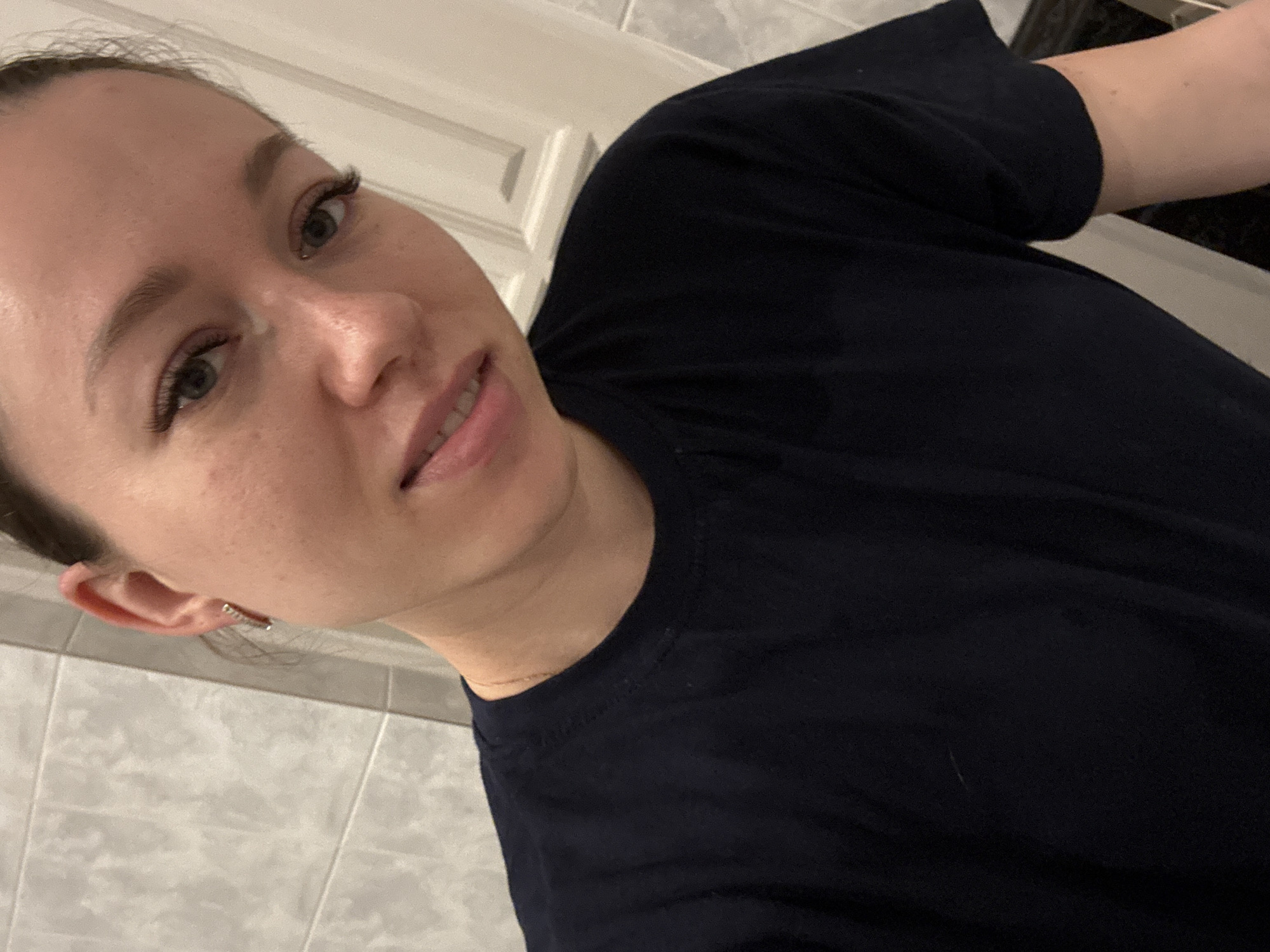 A selfie of a woman with fair skin, light brown hair pulled back from her face, green eyes, and wearing a dark blue T-shirt. She is positioned in what appears to be a bathroom with light gray tiled walls and a white door or cabinet in the background. The woman is looking directly at the camera with a slight smile. She has eyelash extensions and is wearing a small earring in her left ear. A gold chain is visible at her neck. No specific details indicate the location within Ферма, Russia.
