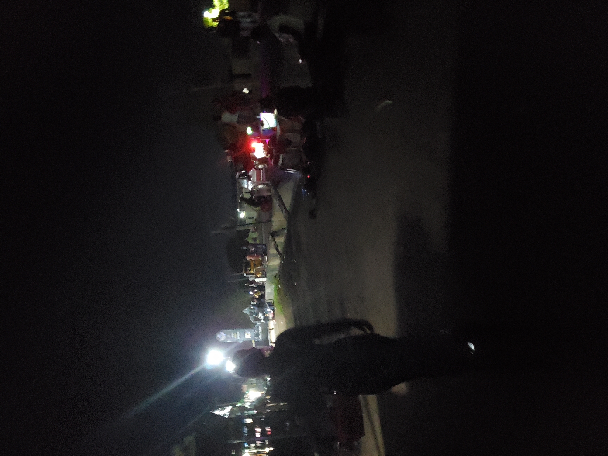The image captures a nocturnal scene in Jalingo, Nigeria, likely an outdoor market or gathering area. In the foreground, a silhouetted individual walks away from the camera on a paved surface, possibly a road or walkway. To the left, structures with illuminated signs suggest commercial activity, with some light reflecting off them. The background reveals a cluster of people and vehicles, including what appears to be a red vehicle with its brake lights illuminated. There's evidence of activity around this vehicle, with some people standing and others possibly seated. Lights from streetlamps or nearby buildings illuminate the scene, creating a low-light environment with distinct light sources and shadows. The overall atmosphere is that of a busy, open-air space after dark.