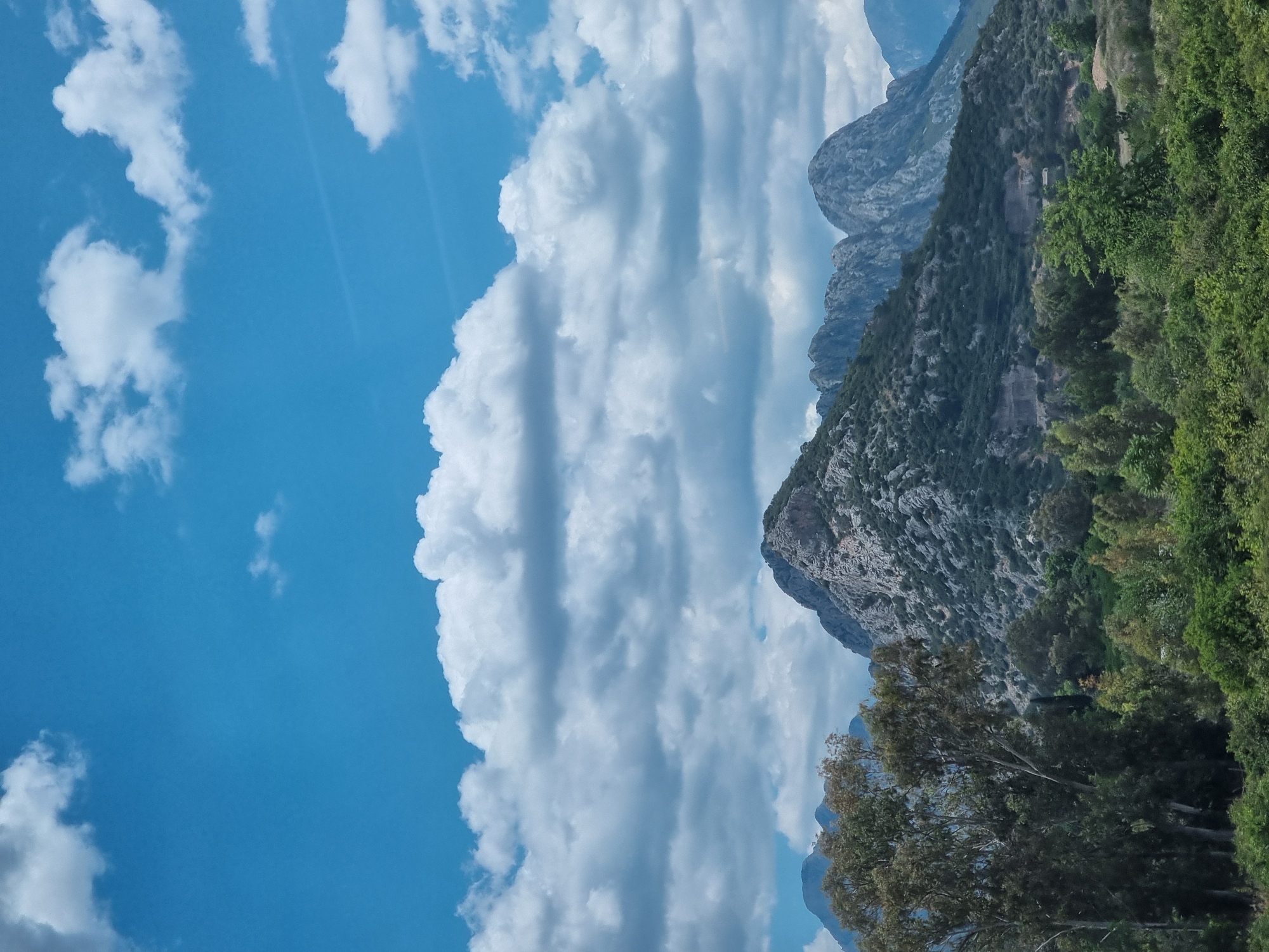 A landscape photograph shows a view in Konyaaltı, Turkey. The sky is mostly blue, with white cumulus clouds, some dense and striated, and others smaller and scattered. On the right side of the frame are mountains; the lower slopes are covered with trees, while the higher elevations are rocky. A few trees are in the foreground.