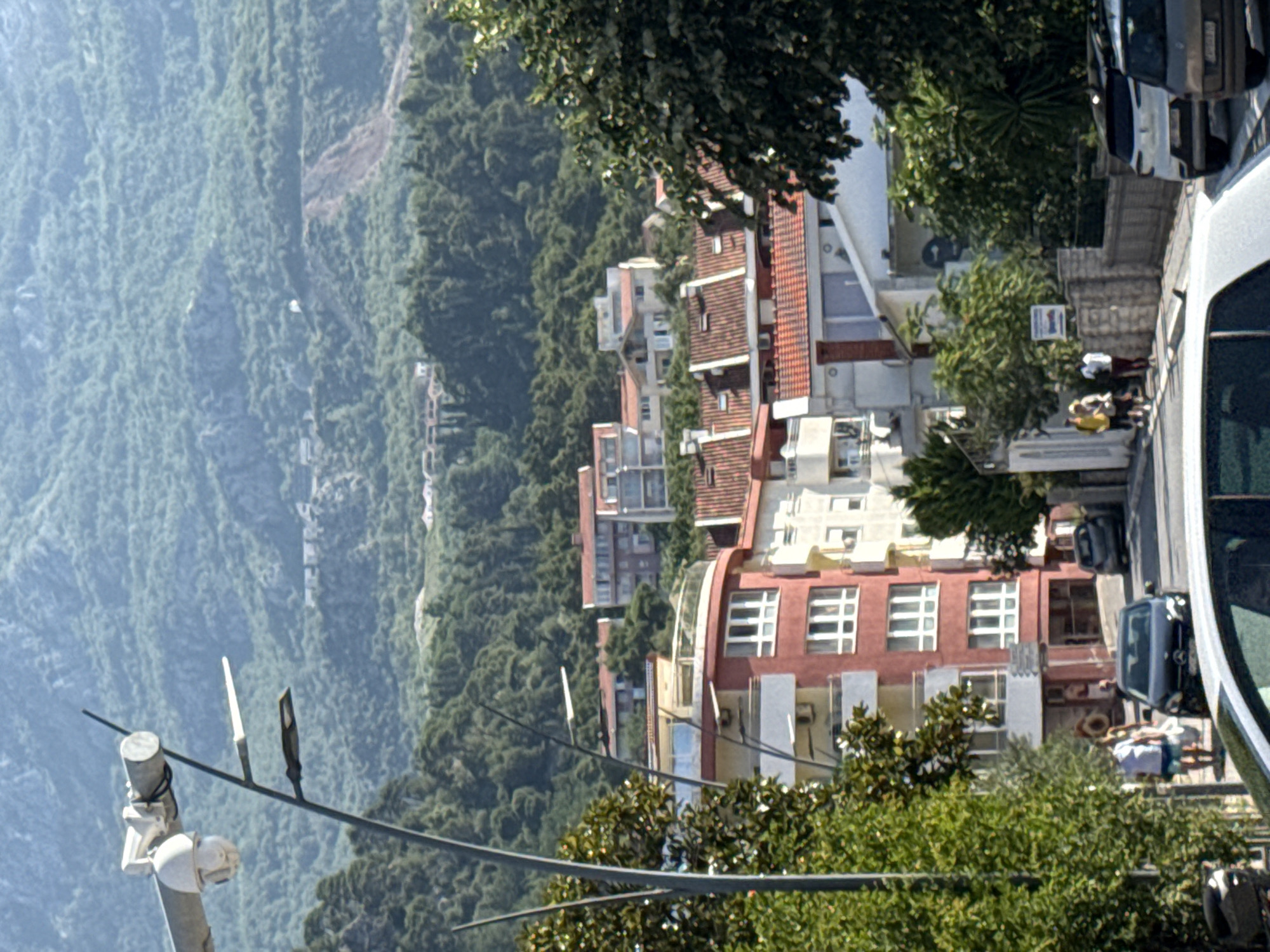 The image shows a street in Budva, Montenegro, with a steep incline. Buildings with red-tiled roofs are visible on the hillside, interspersed with green trees and vegetation. A security camera and a street lamp are mounted on a pole in the foreground. Several cars are parked on the street, and a few people are walking on the sidewalk. In the background, a lush green mountain landscape dominates the scene. The architecture of the buildings suggests a Mediterranean style, and the overall atmosphere is one of a sunny, urban environment nestled within nature.