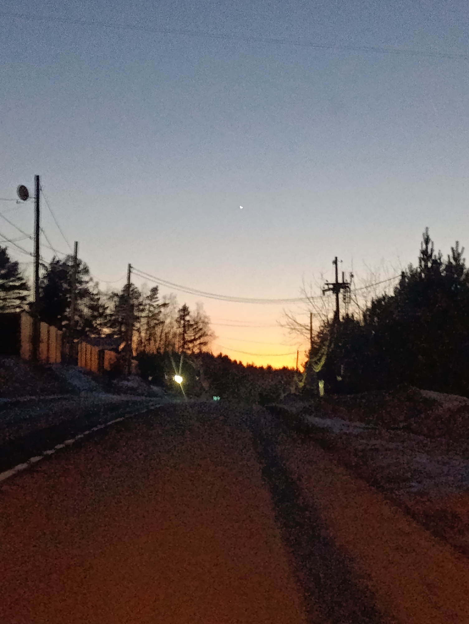The image depicts a rural road at twilight, likely a back road within or on the outskirts of Krasnoyarsk, Russia, given the presence of utility poles and the style of the fencing. The sky is a gradient of blue to orange, indicating either sunrise or sunset. A sliver of the moon is visible in the upper center of the frame. The road surface is rough and appears to be asphalt or gravel, with patches of snow or frost along the edges. The scene is bordered by trees and shrubbery on both sides, and the light from a distant street lamp or vehicle is seen further down the road. The perspective is from ground level, as if the photo was taken from within or very close to the road. There is no visible human activity in the immediate area.