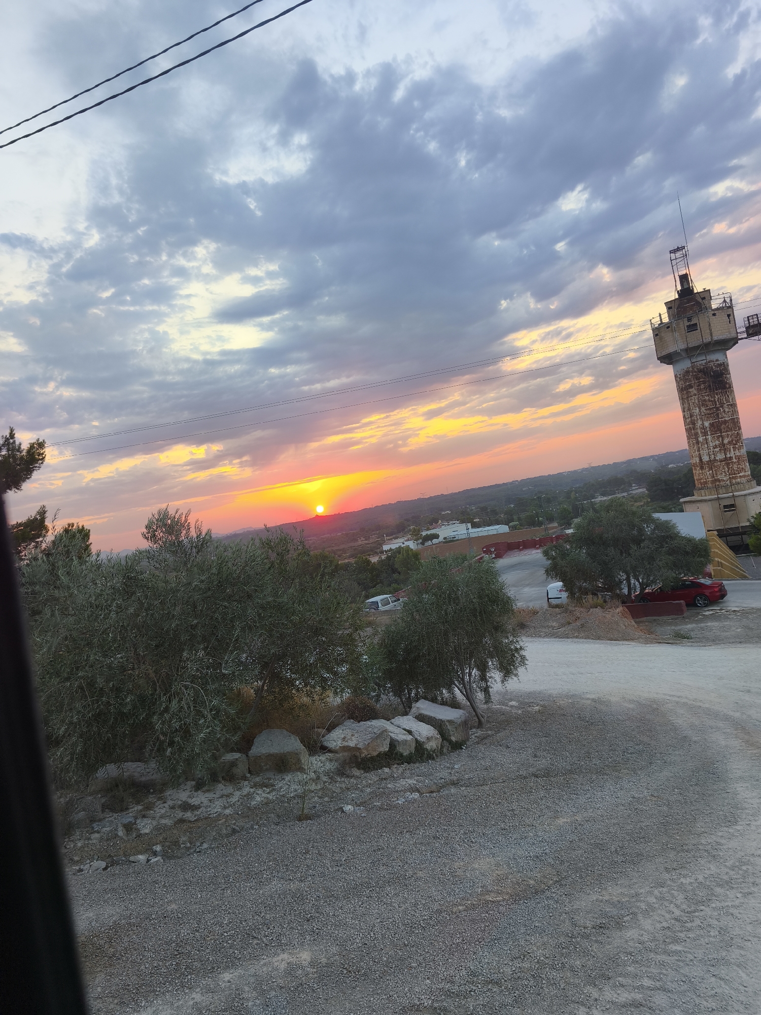 The image depicts a sunset scene in Riba-roja de Túria, Spain. The sky is filled with dramatic clouds, displaying a vibrant gradient of orange, yellow, and pink hues as the sun sets. A tall, rustic water tower stands prominently on the right side of the frame, its weathered facade contrasting with the colorful sky. In the foreground, several olive trees are visible, with their green foliage adding a touch of nature to the landscape. The ground is covered with gravel, and a few vehicles, including a red car and a white van, are parked nearby. The overall impression is one of a rural or semi-industrial area experiencing the beauty of a sunset.