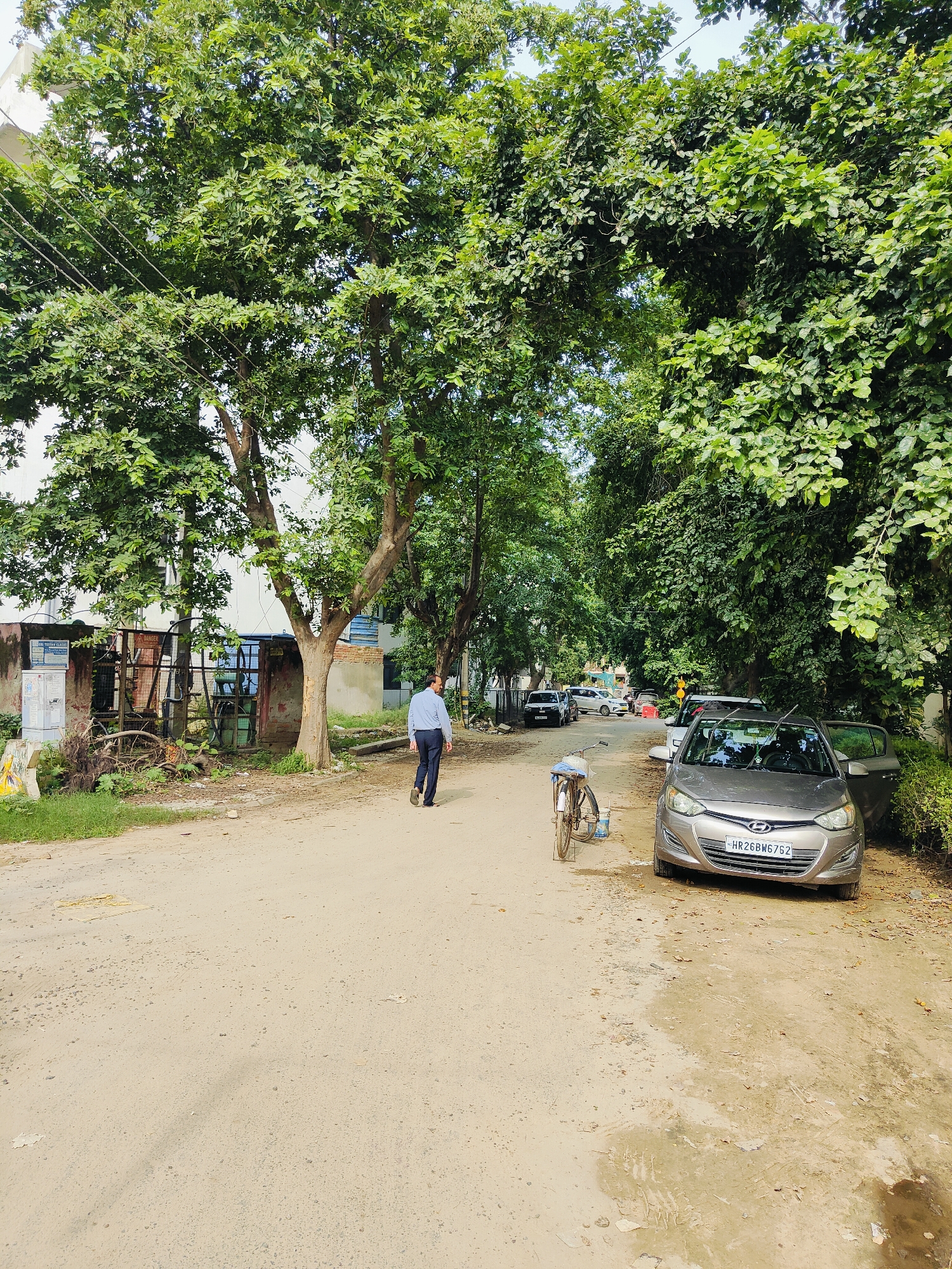 A dusty, unpaved road passes through a tree-lined area in Gurgaon, India. On the left side of the road, there is an industrial-looking structure with a metal fence and what appears to be electrical equipment. A man in a light blue shirt and dark trousers walks away from the camera down the road. To the right of the road, a light gray car is parked, its passenger door open. A bicycle with a basket is leaning against the car. Further down the road, several other cars are visible. Lush green trees with dense foliage overhang the road, creating a shaded environment. The sky is visible through the canopy of leaves.