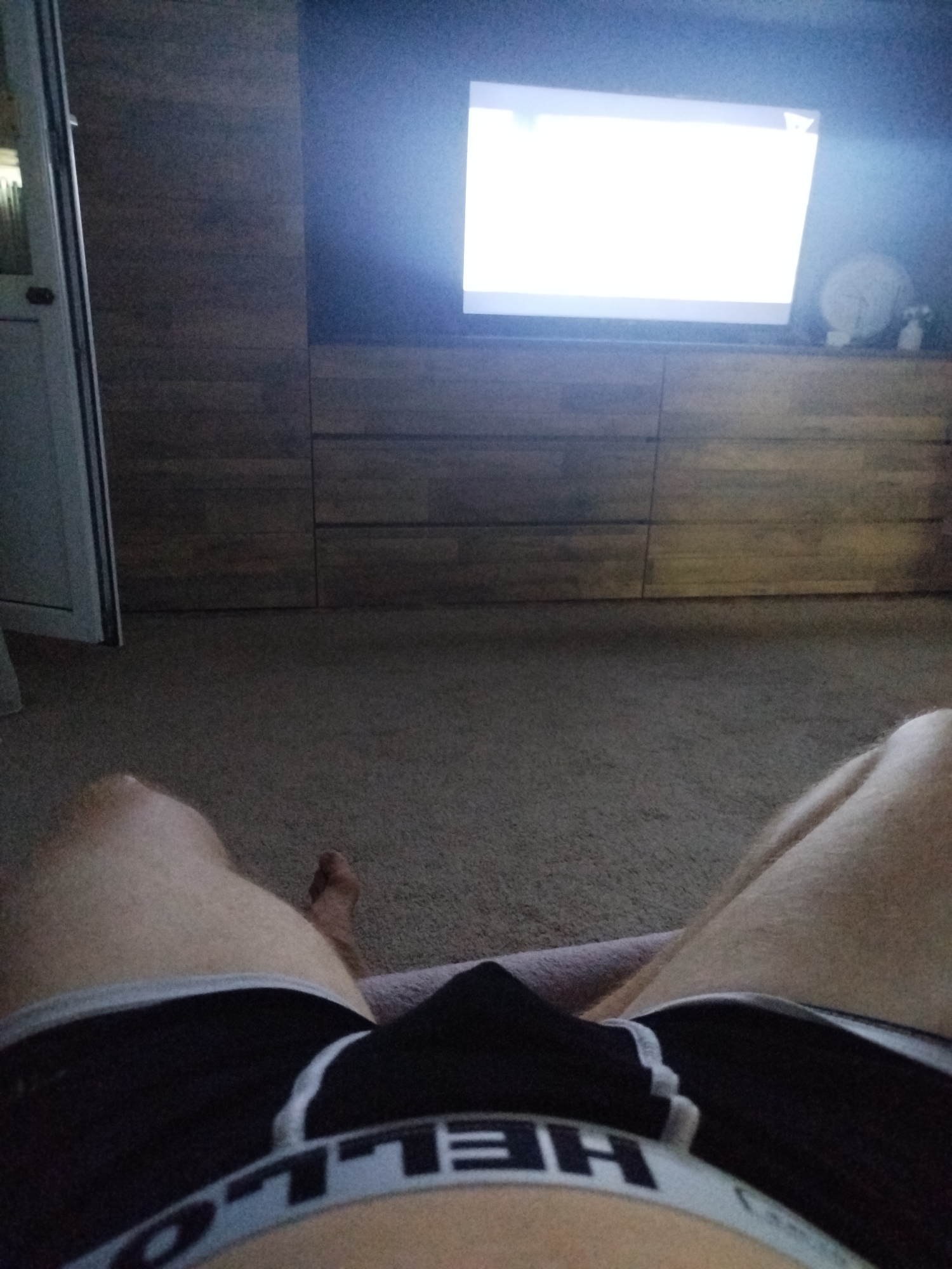 The image shows a first-person perspective of someone lying down in a dimly lit room, with a large television screen illuminated in the background. The person is wearing black underwear with a white waistband that has lettering on it. The room appears to be a living space with wooden furniture. The floor is carpeted. There are no visible indications of the specific location within Naberezhnye Chelny, Russia.