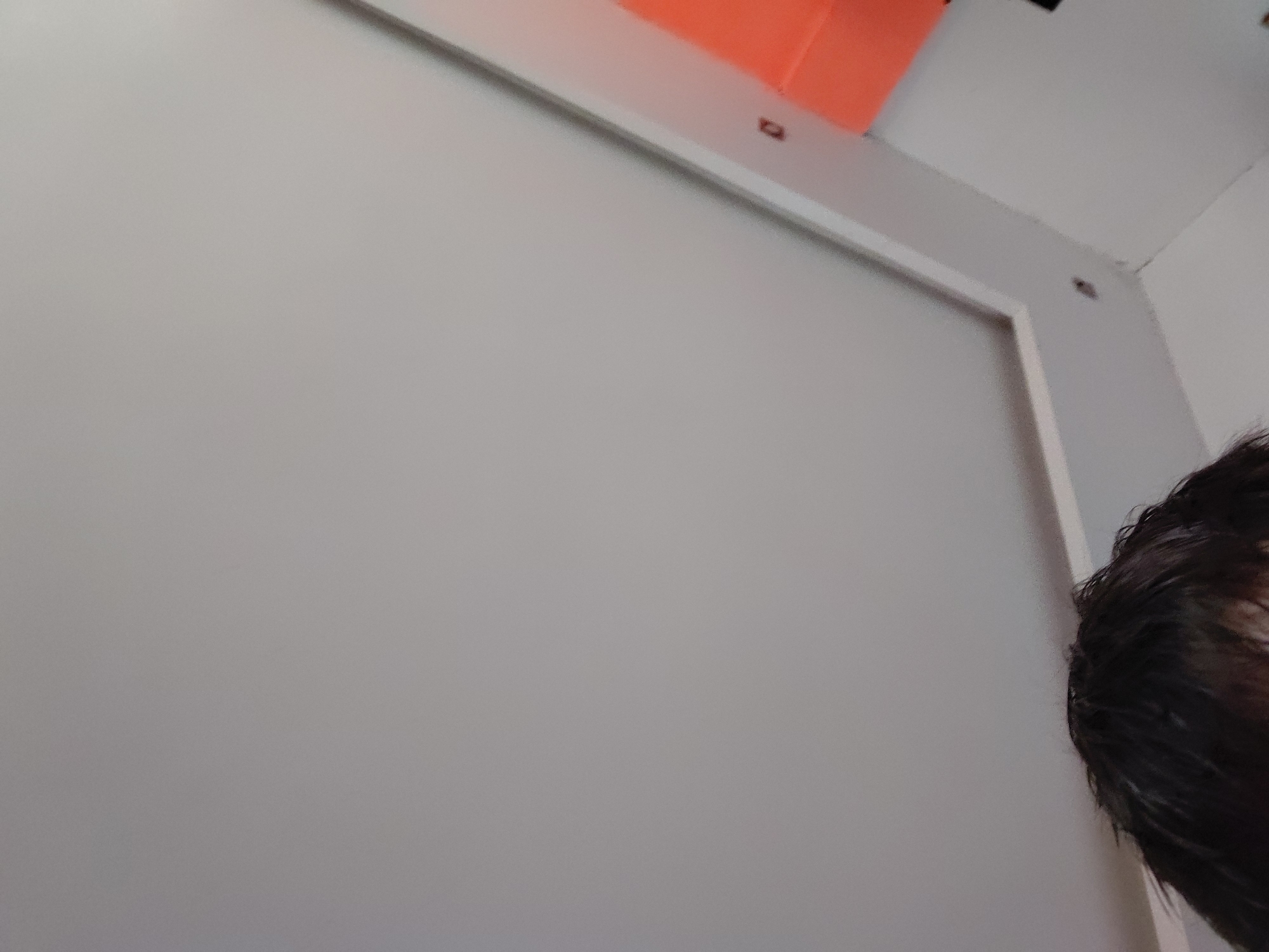 The image captures an indoor scene, primarily featuring a large, plain light gray wall or panel that dominates the left and center of the frame. A white architectural trim or frame runs horizontally along the top edge of this gray surface and vertically down its right side.
In the upper left corner, partially obscured by the trim, is a bright orange object, possibly a box, fixture, or part of a larger structure. Above the white trim, towards the top right, a corner of a white ceiling or upper wall is visible, suggesting the room's boundary. There are a couple of small, dark, circular elements embedded in or attached to the white trim and the wall above it, which could be screw heads, mounting points, or small decorative accents.
On the far right side of the image, the top and side of a person's head with dark, possibly wet or styled, hair is visible, cropped tightly at the edge of the frame. The lighting appears even, suggesting an interior space during the day or under artificial illumination. No specific activity is depicted, and no text is visible. The scene appears to be a static shot of an interior within Santa Ana, Venezuela, but there are no specific visual cues to confirm the location beyond the general architectural elements.