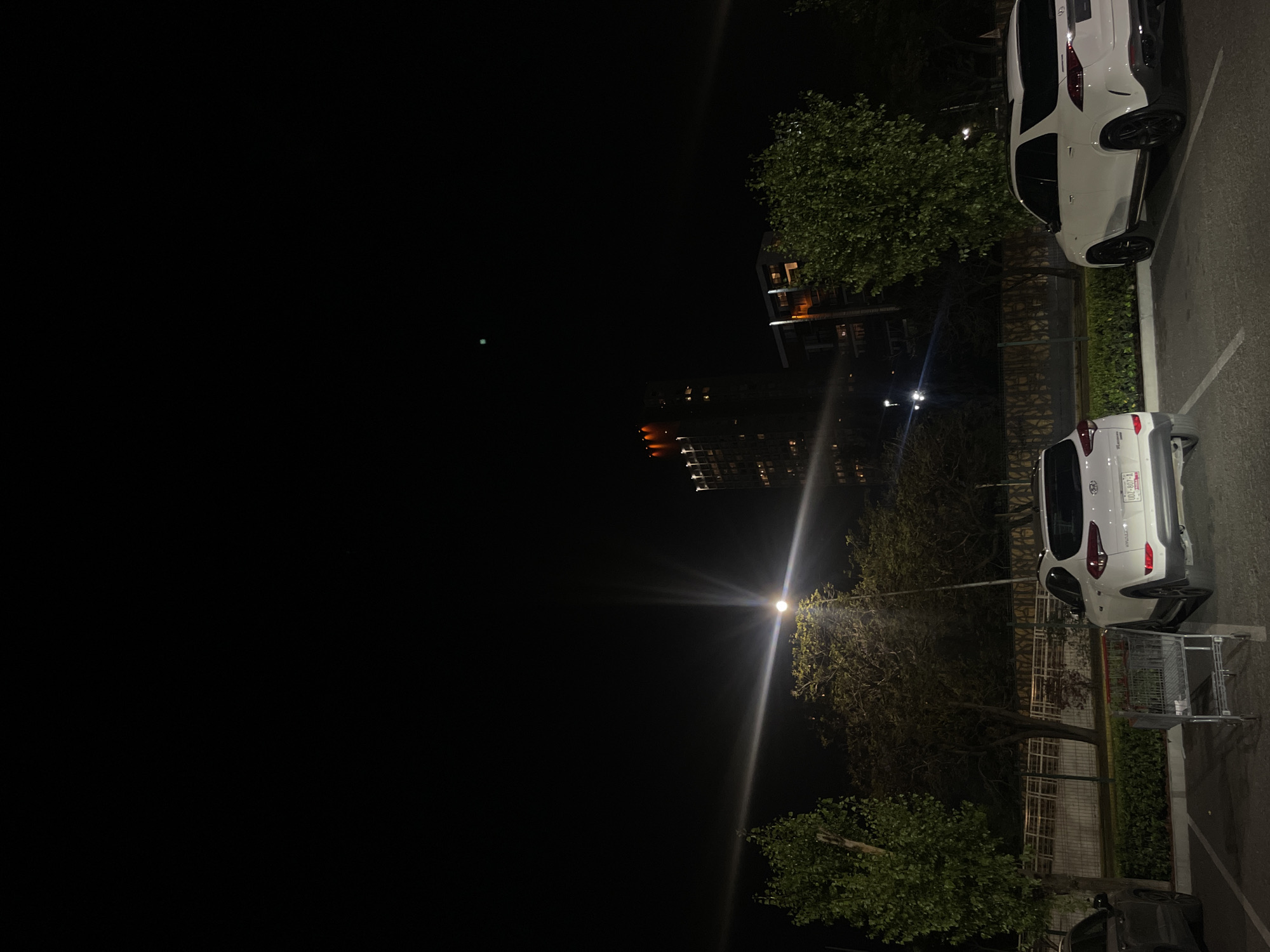 At night in Puebla, Mexico, the photo depicts a parking lot scene illuminated by a street light. Two parked white SUVs, a shopping cart, trees, buildings in the distance, and a tall structure are visible.