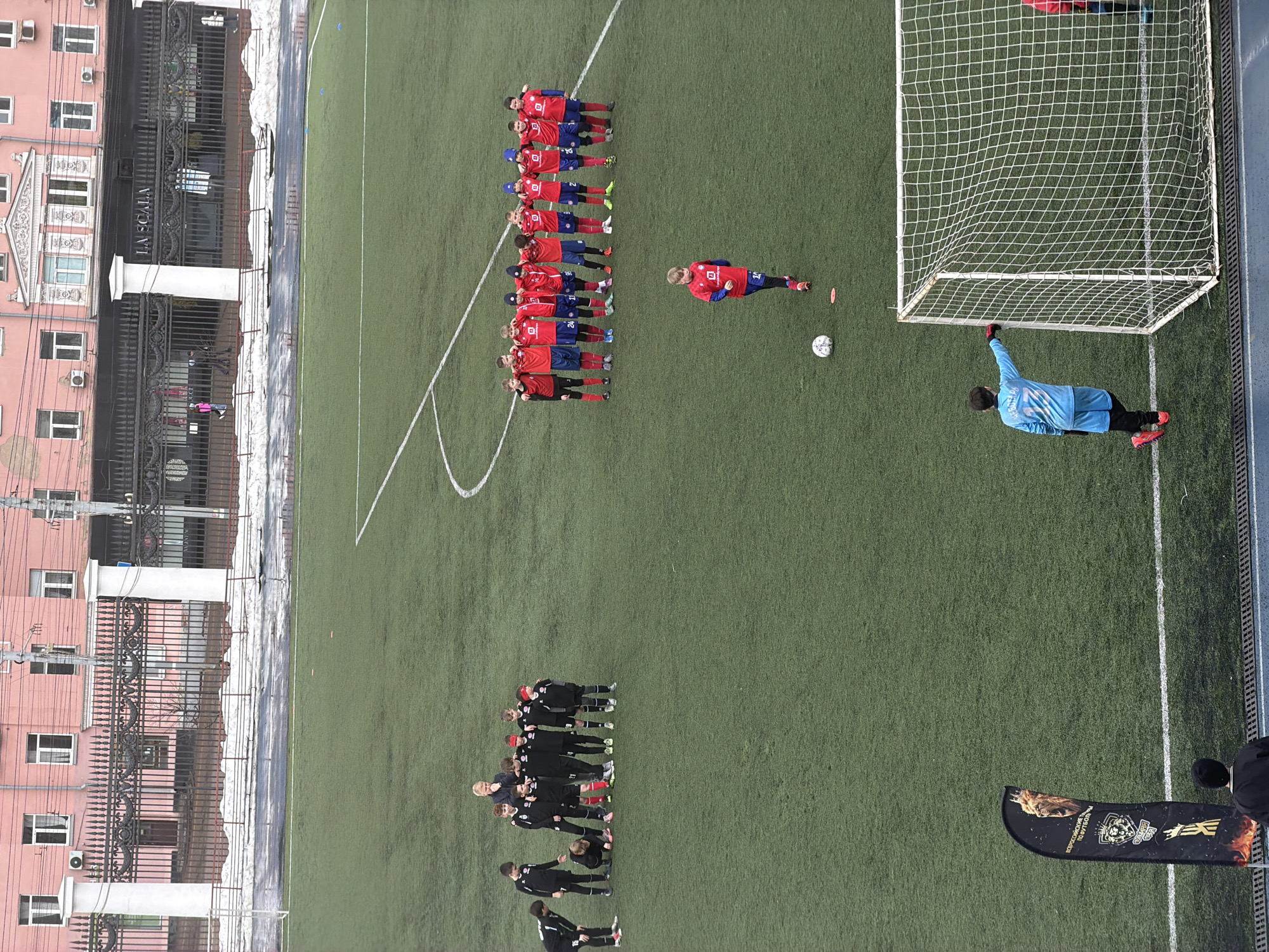 The image shows a soccer field, likely artificial turf, with two teams. One team, dressed in red and blue, is lined up along the edge of the penalty box near a soccer ball and the goal. A single player in red and blue is slightly removed from this line and is also near the penalty spot. The goalkeeper, dressed in light blue, is reaching to block a possible shot. Another team, dressed in black, is lined up further down the field. The location includes buildings with "La Scala" written on them and a soccer flag. The image was taken in Izhevsk, Russia.